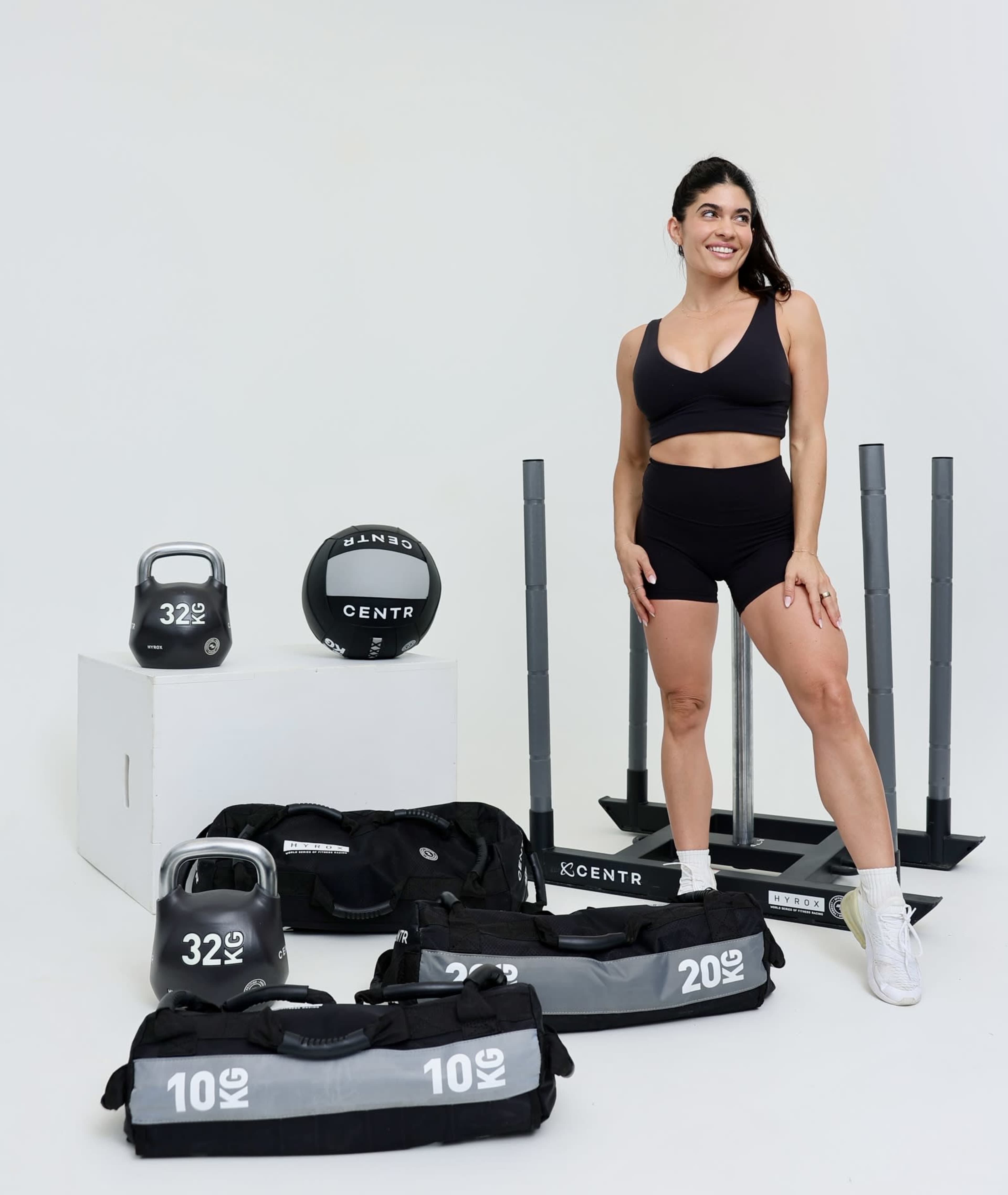 A woman in athletic wear stands beside various gym equipment, including kettlebells, weights, and a medicine ball, arranged on a plain background.