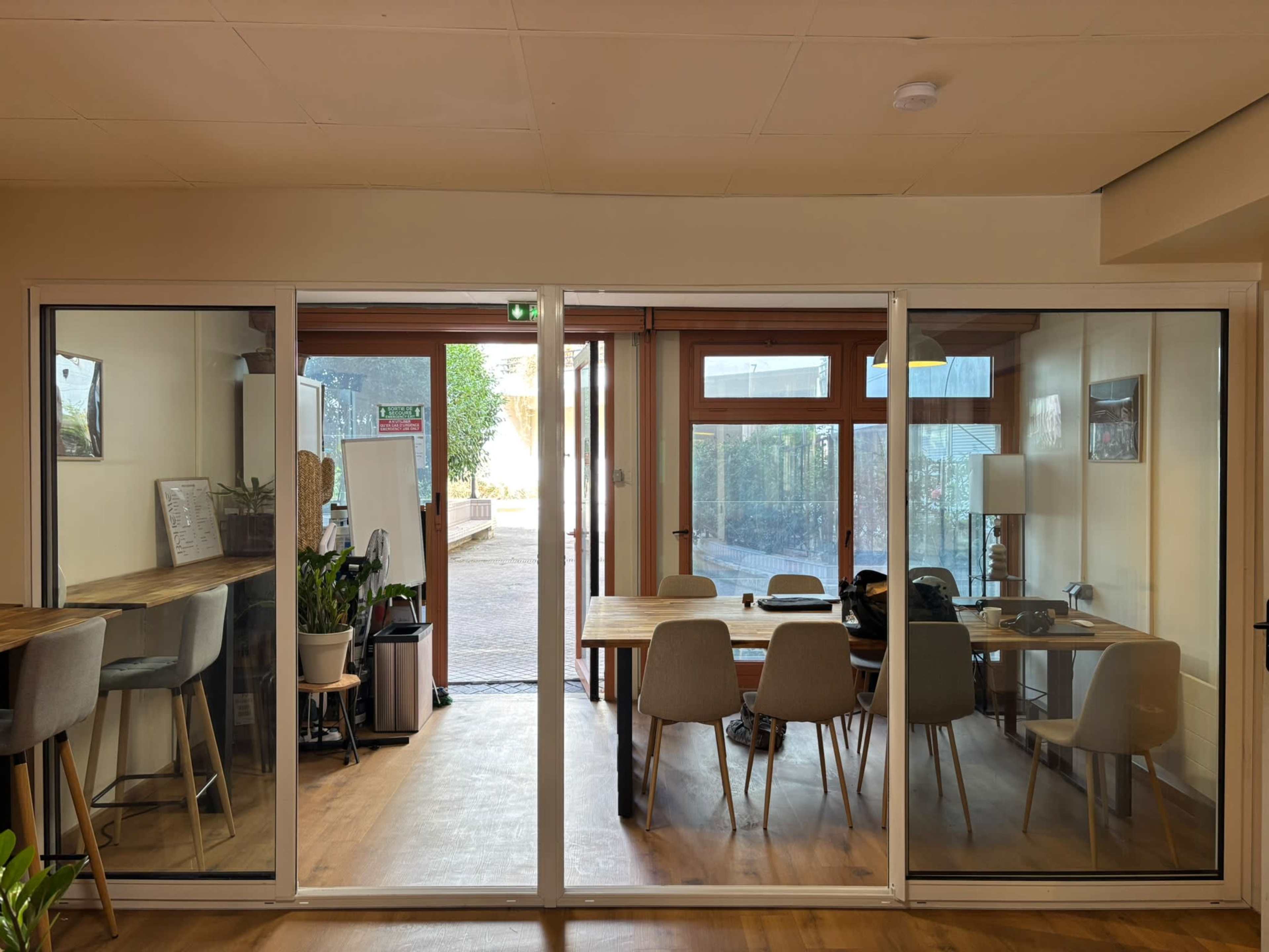 The image shows a spacious, well-lit room with large glass doors leading to an outdoor area, featuring a dining table with chairs, a small table, and some plants.