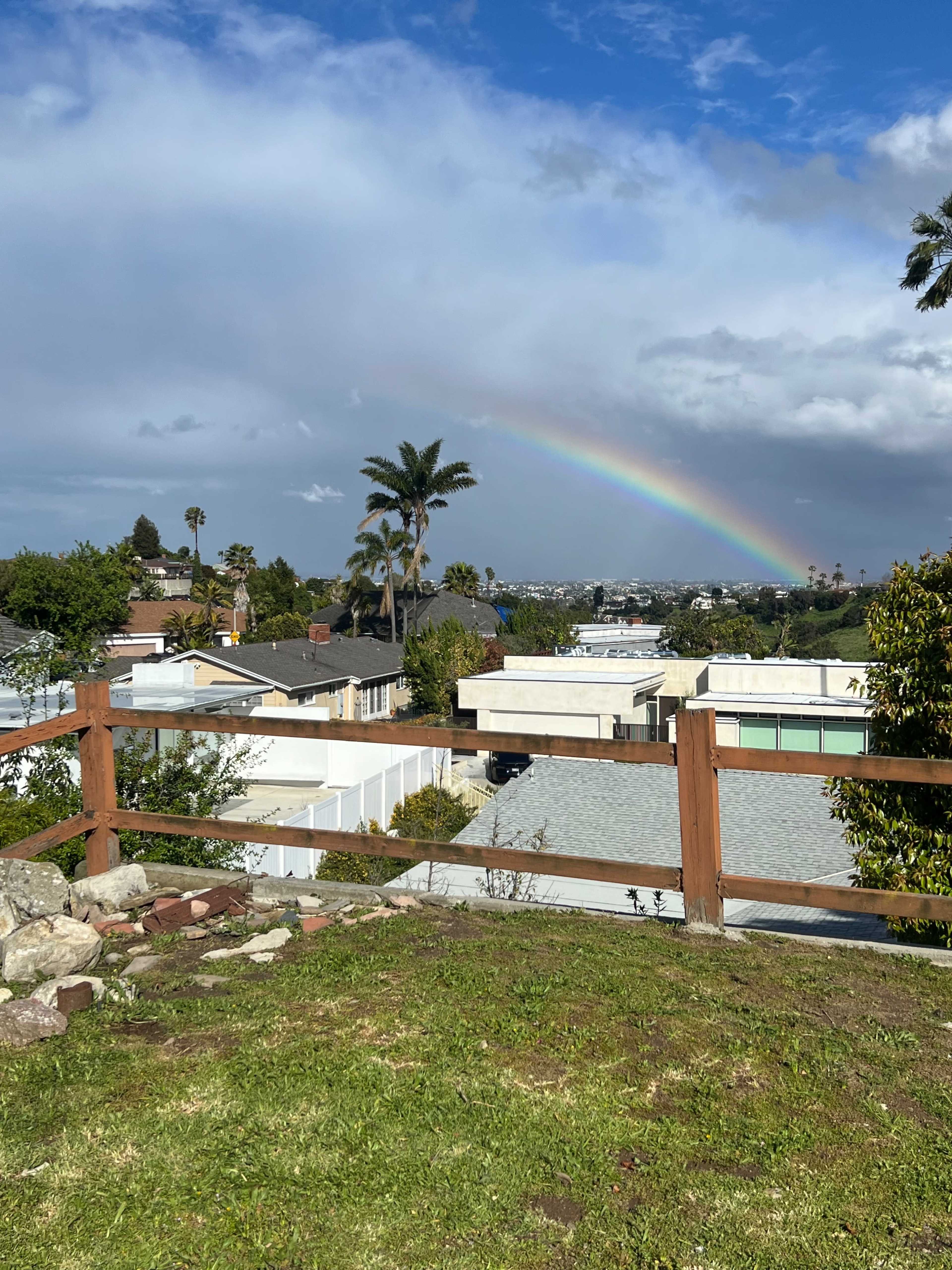 A rainbow arches over a neighborhood with palm trees and houses visible beneath a partially cloudy sky.