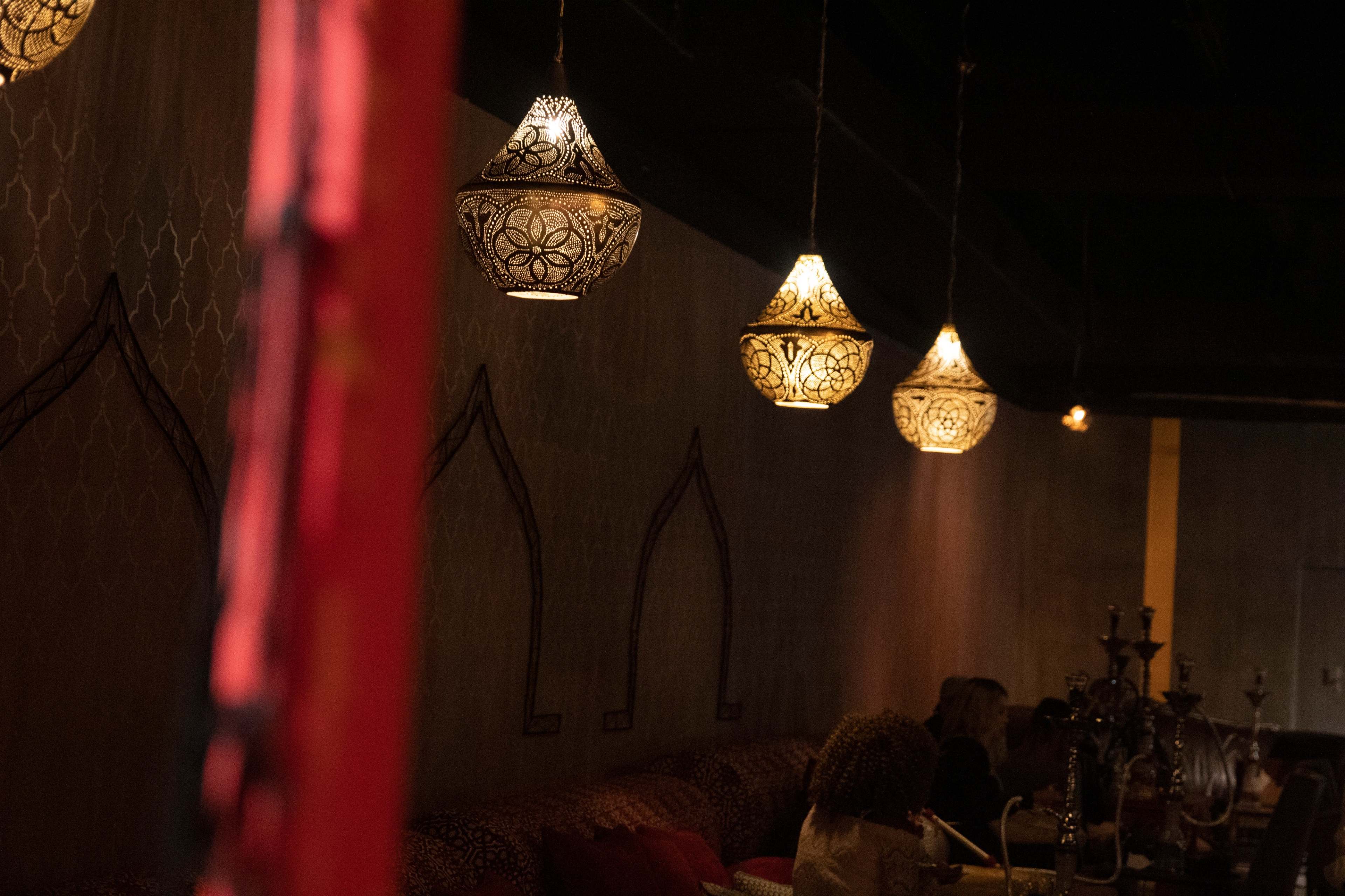 A dimly lit interior of a café or restaurant featuring ornate hanging lamps and a patterned wall with seating along one side.