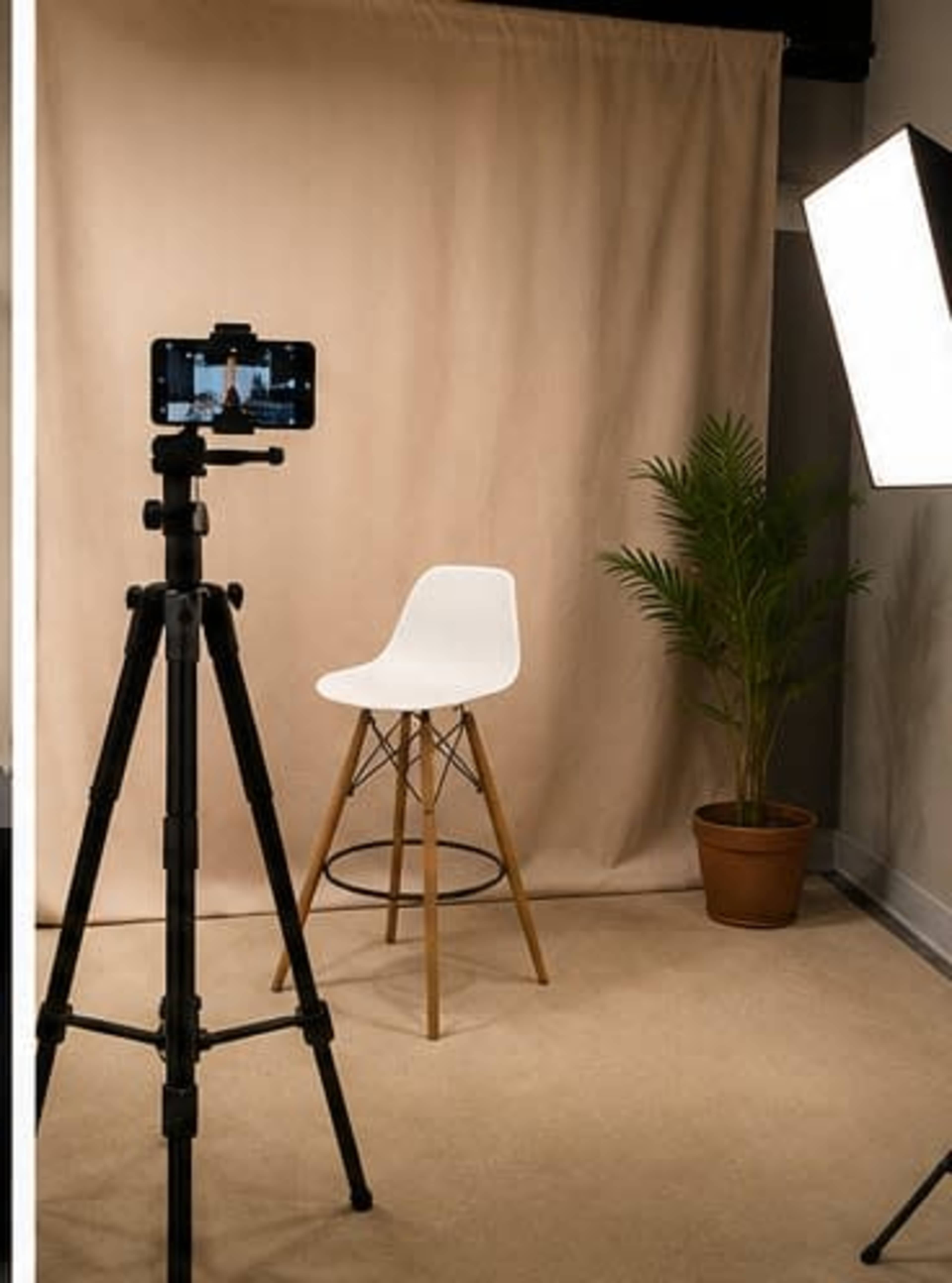 A camera mounted on a tripod faces a white chair in front of a beige backdrop, with a potted plant nearby and a softbox light illuminating the scene.