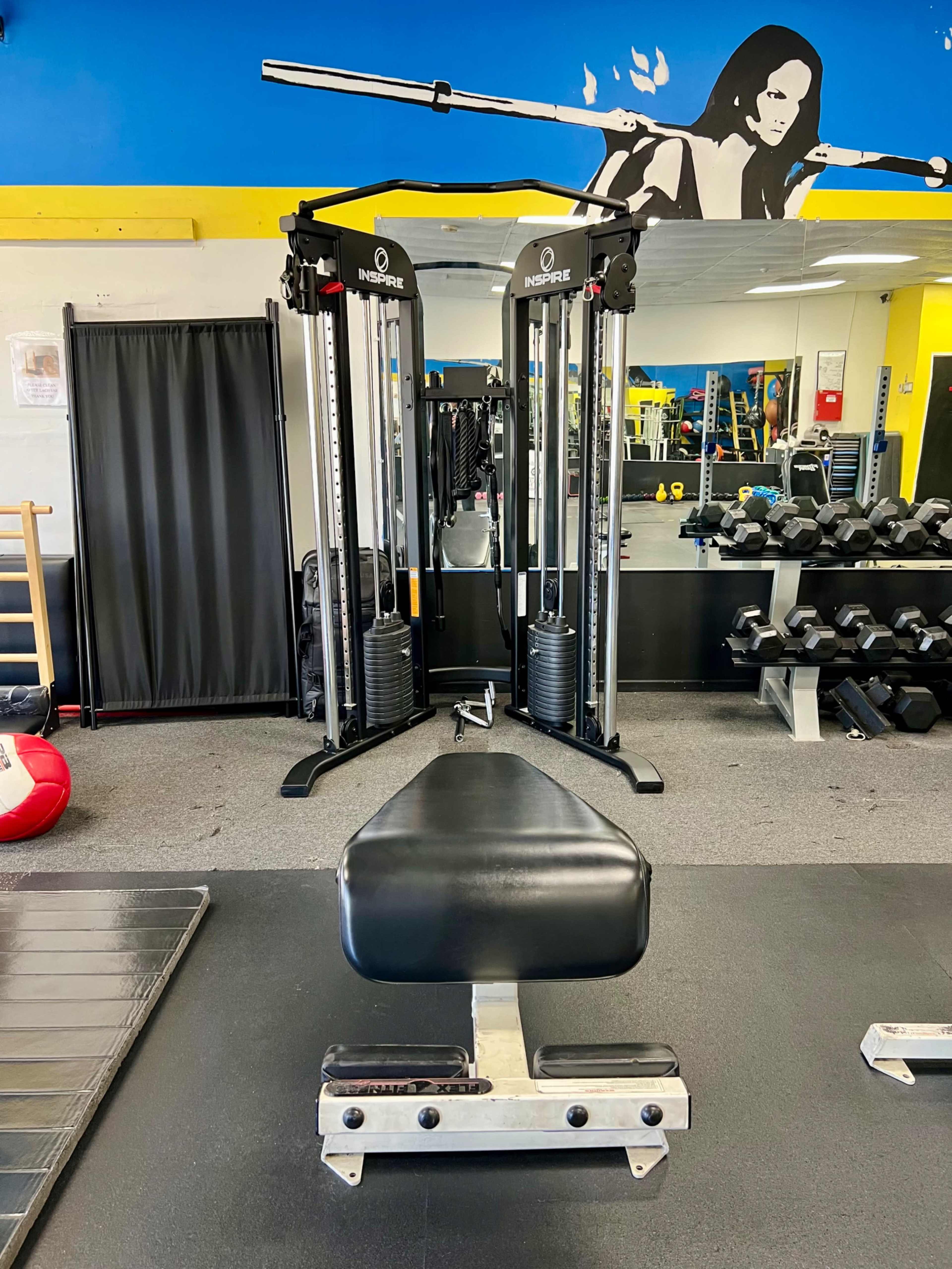 The image shows a gym interior with a dual cable machine in the center, a bench in front, and a variety of weights and equipment in the background.