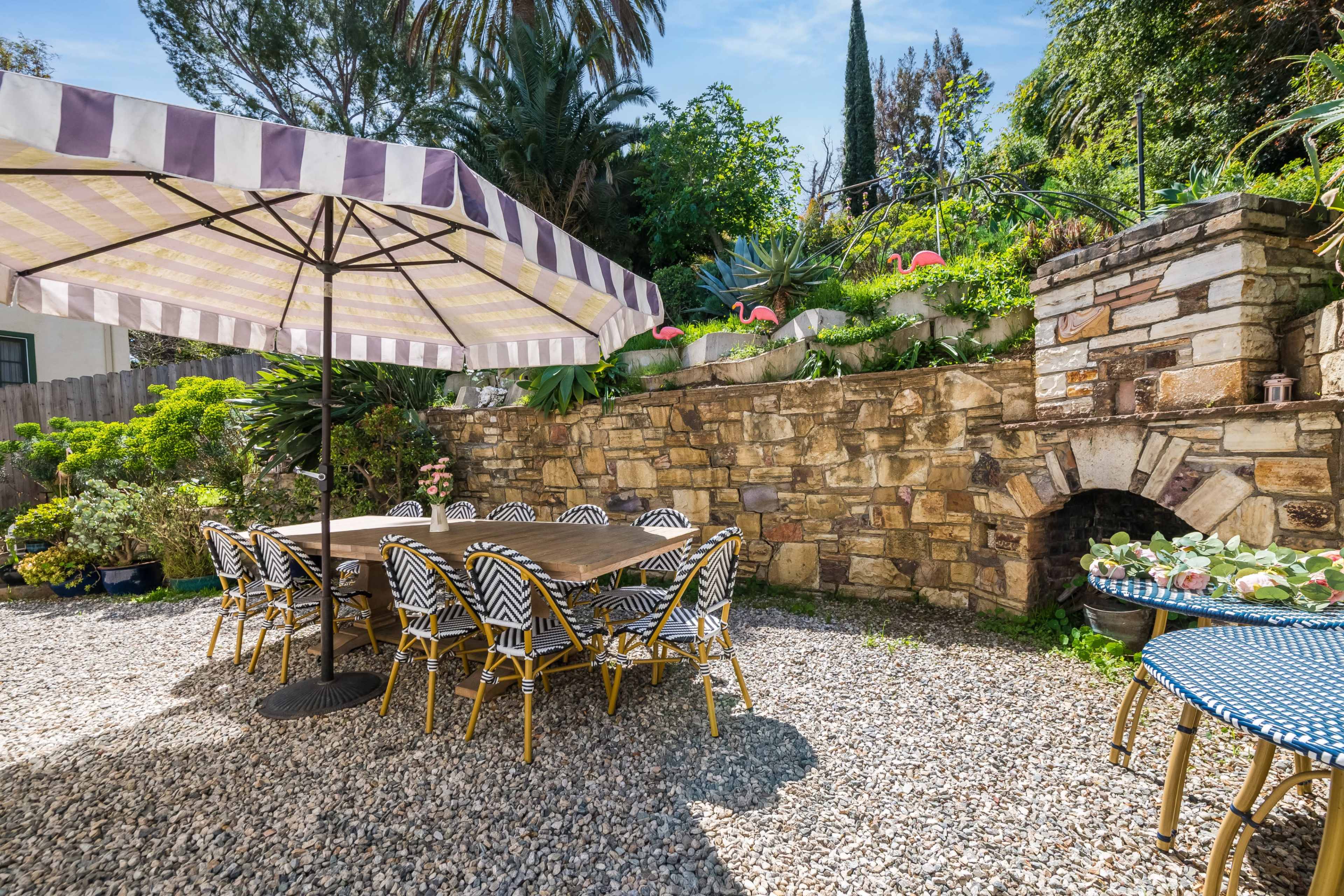A patio area features a large table with striped chairs under an umbrella, surrounded by greenery and a stone wall.