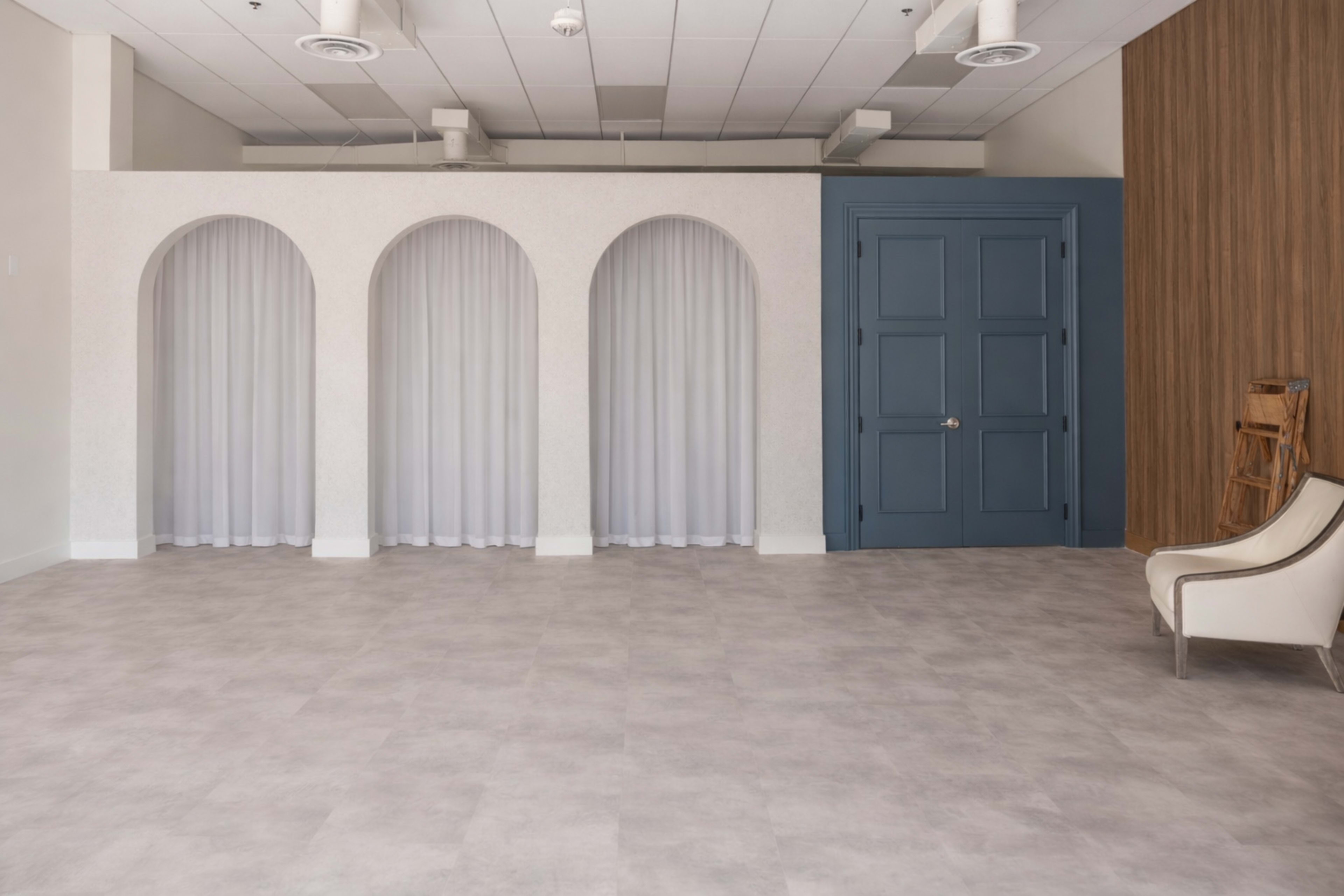 The image shows a spacious room featuring three archways with white curtains and a blue door against a wooden accent wall, along with a light-colored chair in the corner.