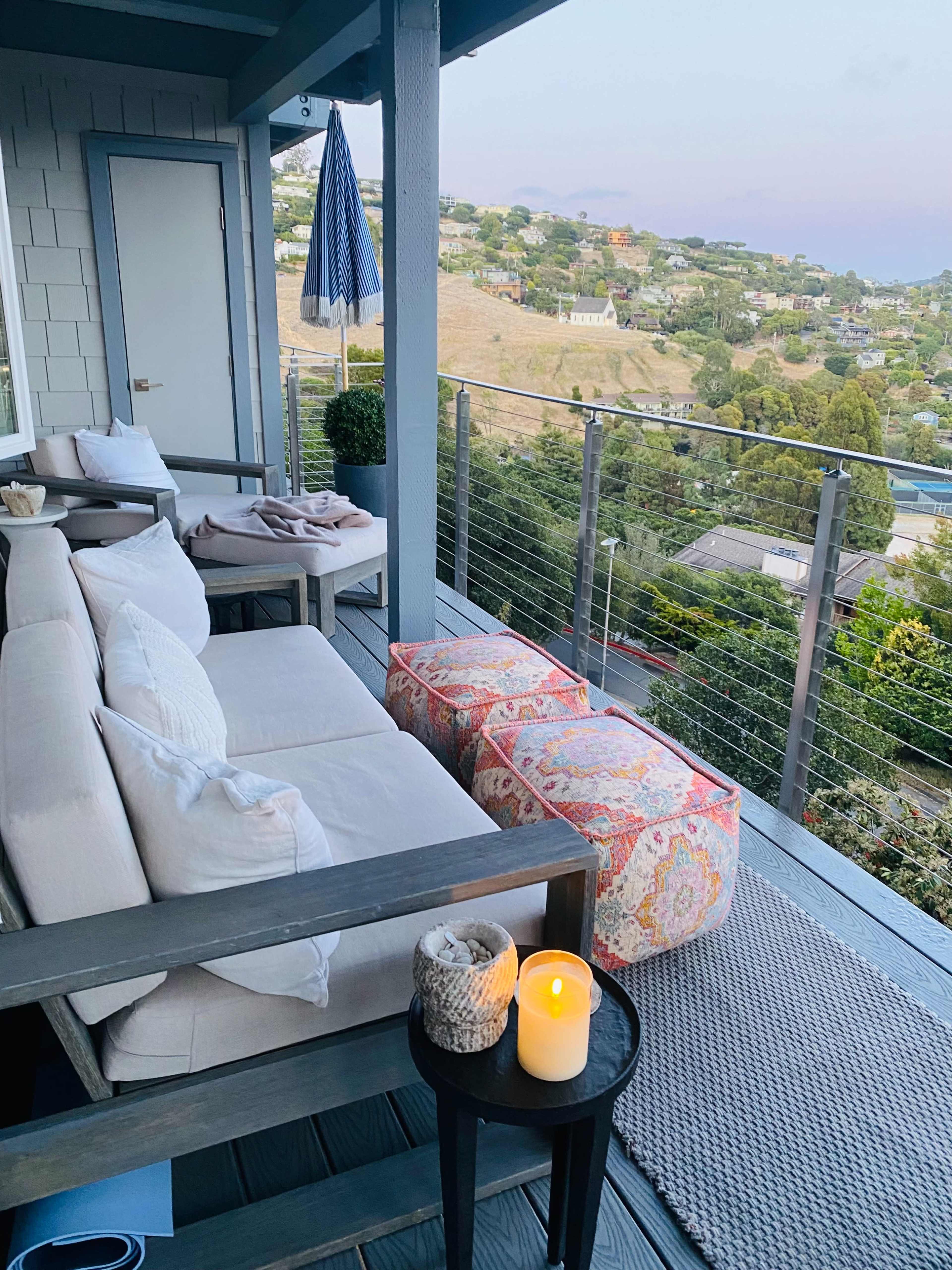 A modern balcony features a seating area with a couch, two decorative poufs, and a lit candle, overlooking a hillside neighborhood.