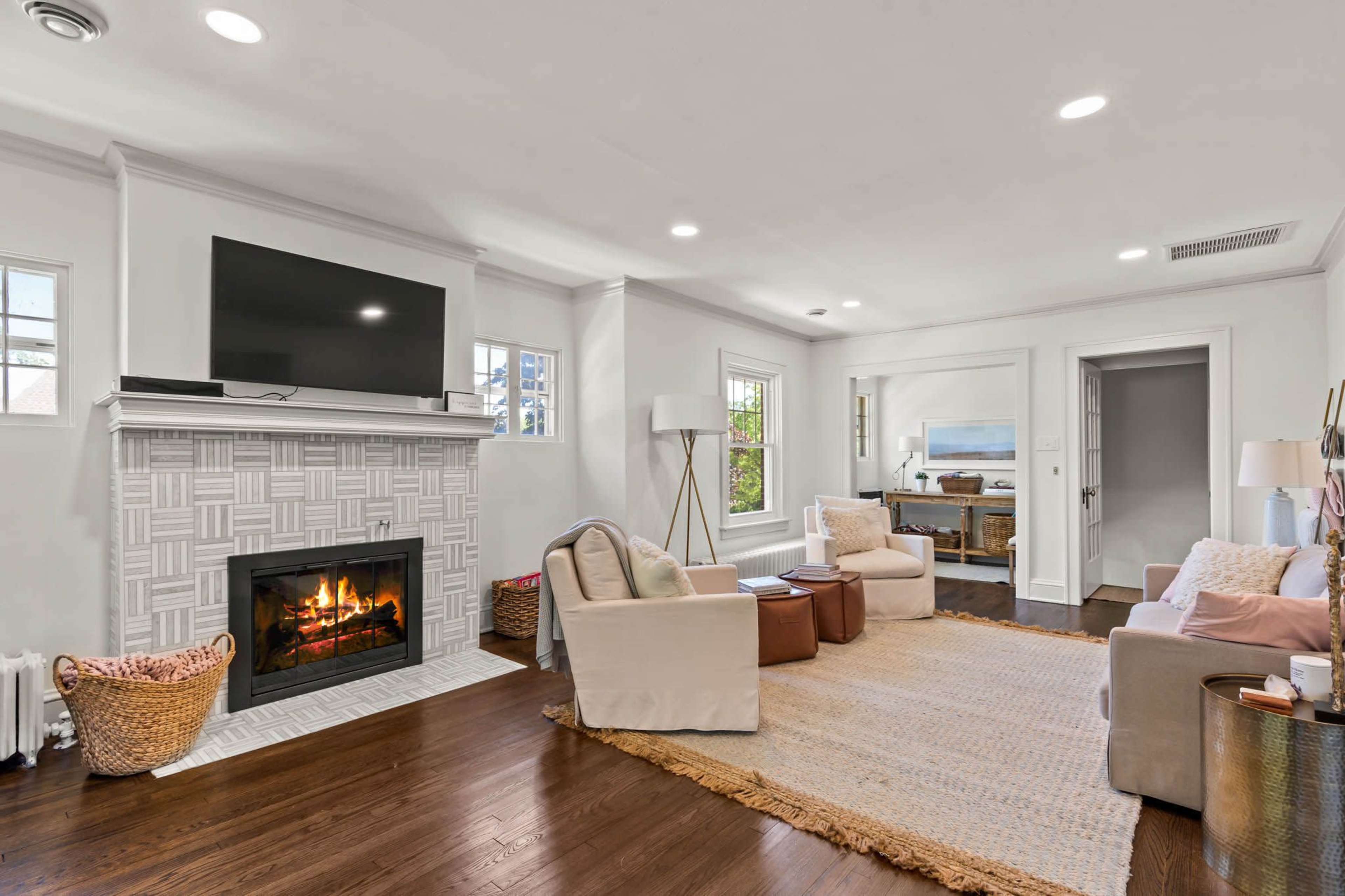 A living room featuring a fireplace with a television mounted above it, two armchairs, a coffee table, and a woven rug on hardwood flooring.