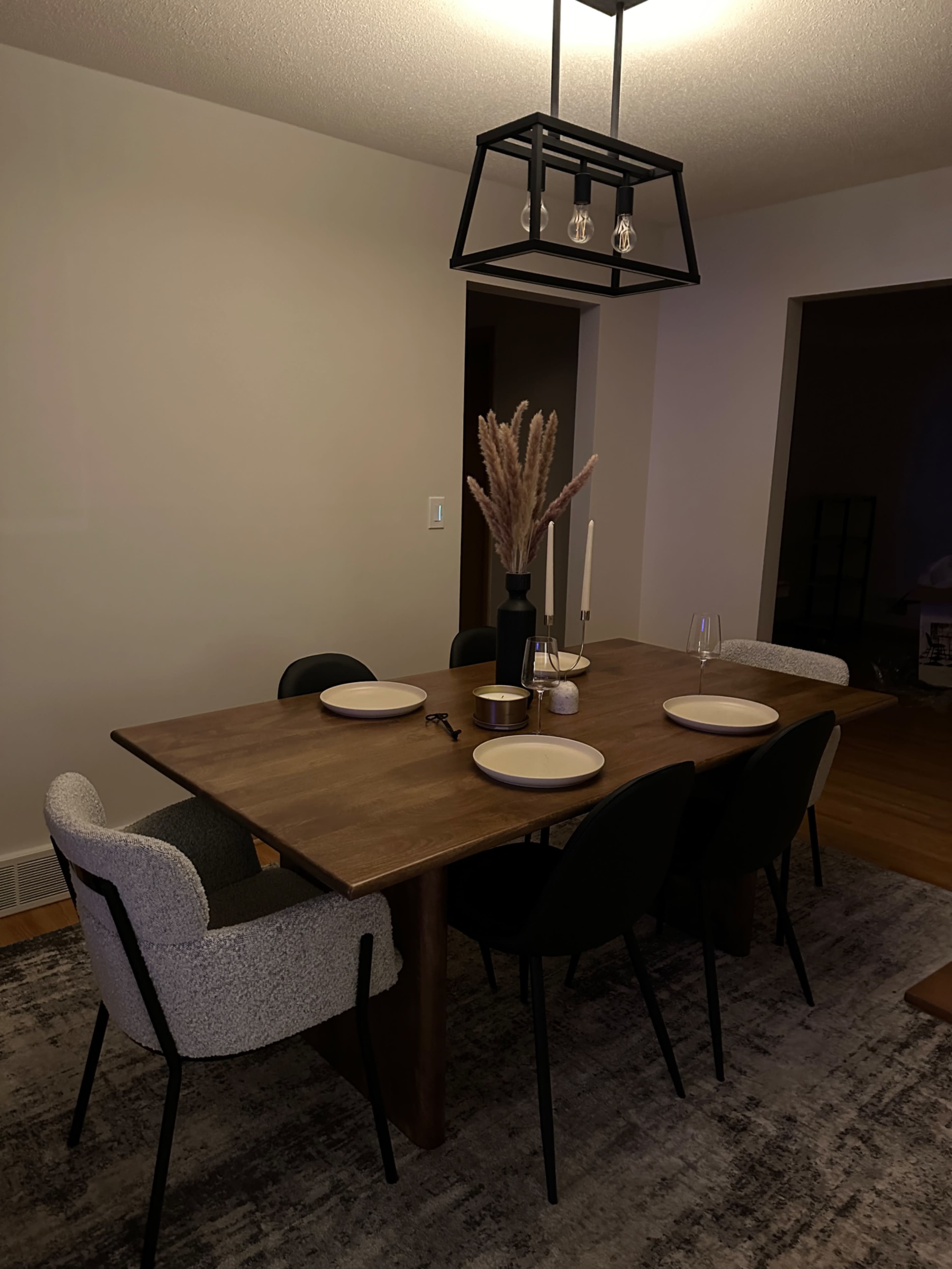 A wooden dining table set for four with plates, candles, and a vase of dried flowers is surrounded by black chairs in a softly lit room.