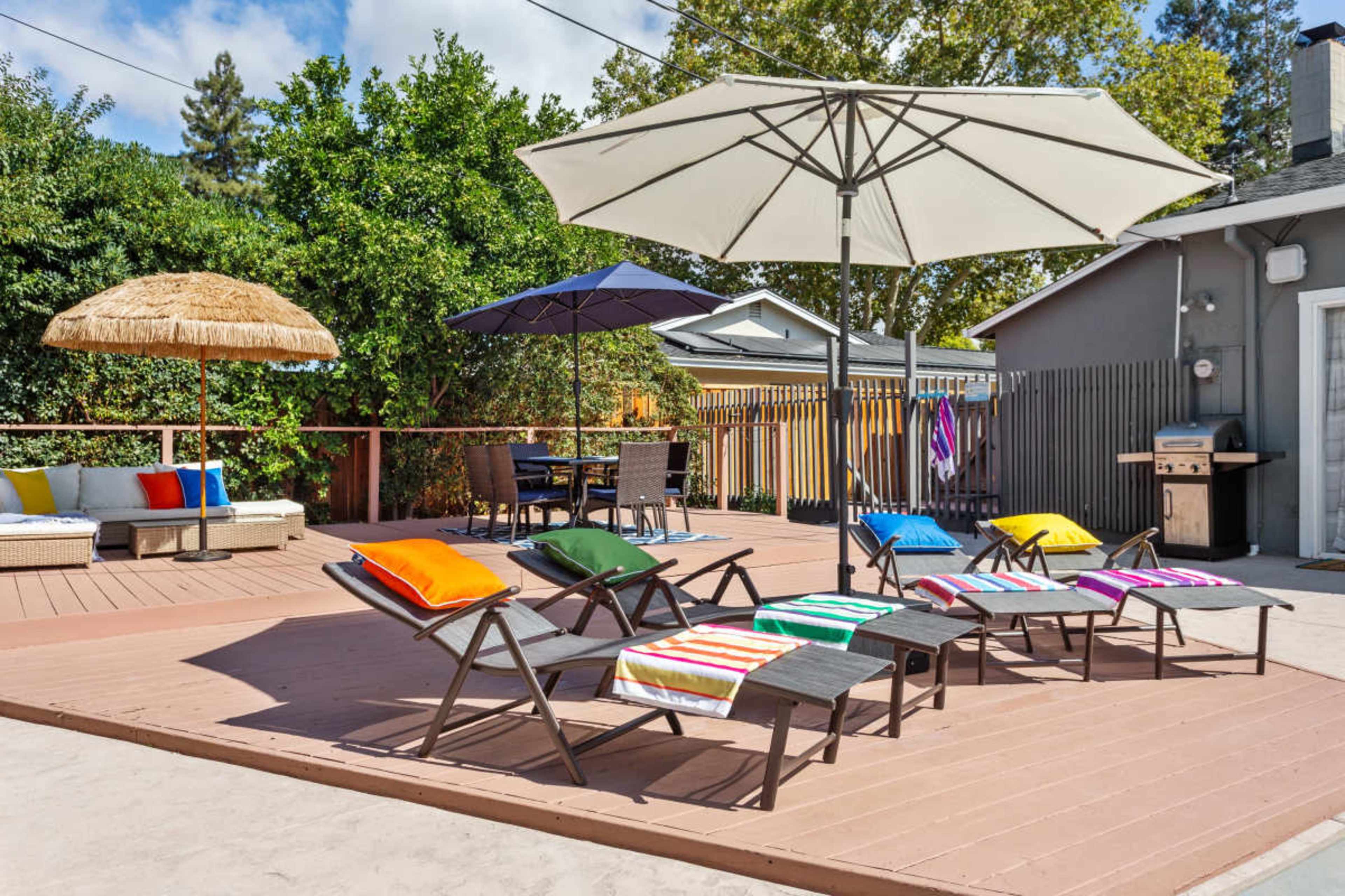 A patio features several lounge chairs with colorful cushions, two umbrellas, and a seating area surrounded by greenery.