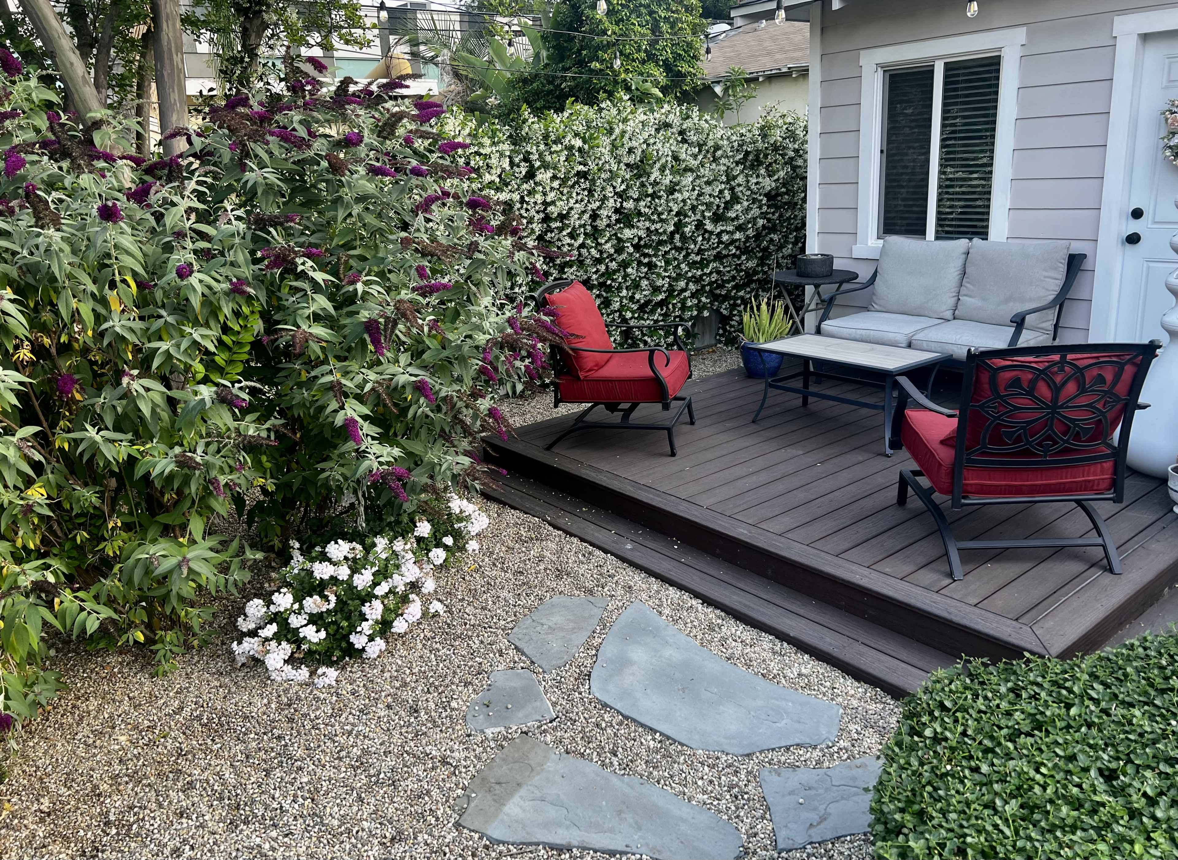 A small outdoor seating area features a wooden deck with two red chairs and a sofa, surrounded by flowering bushes and gravel pathways.