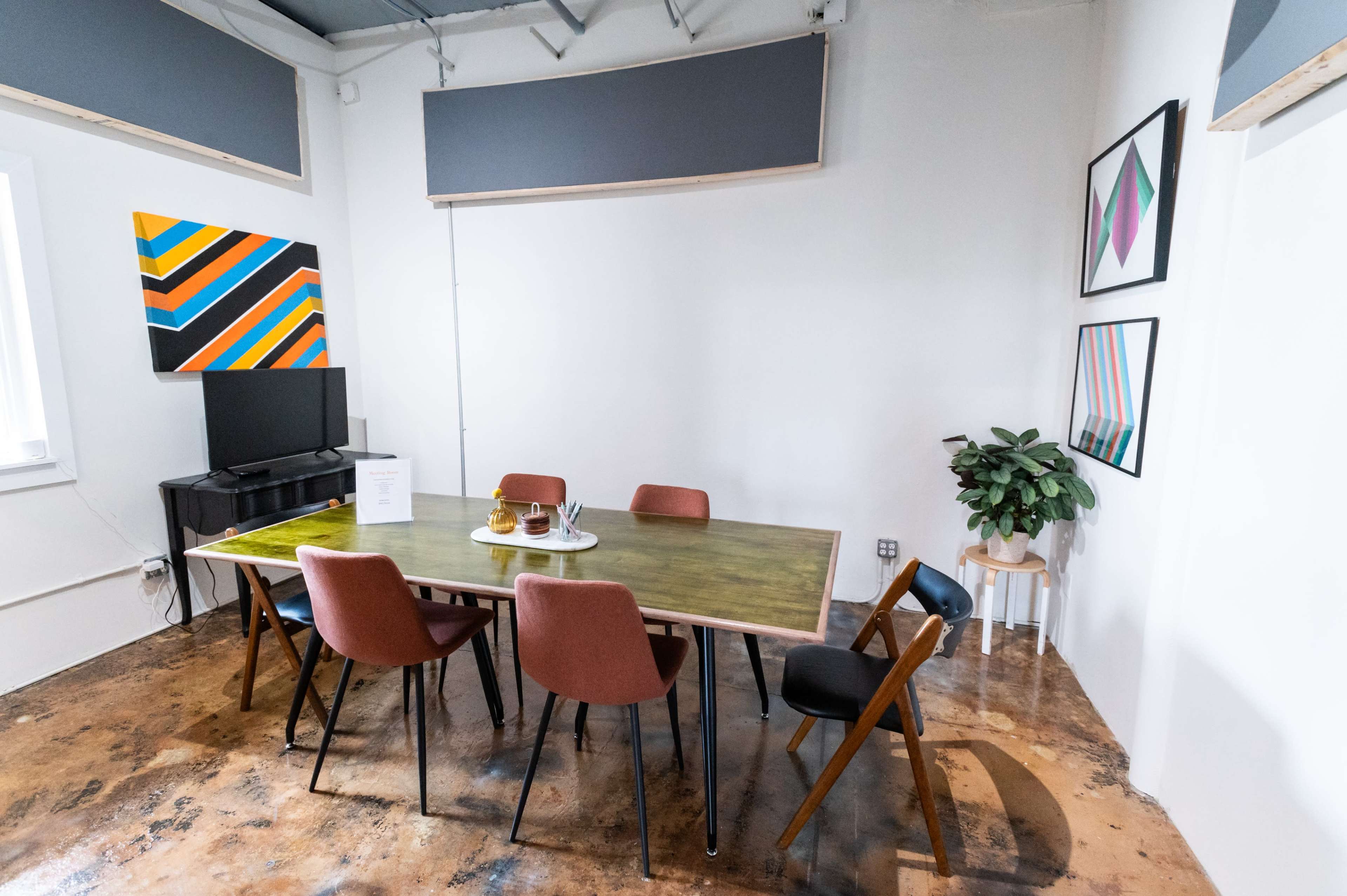 A modern meeting room features a green table surrounded by colorful chairs, a television, and artwork on the walls.