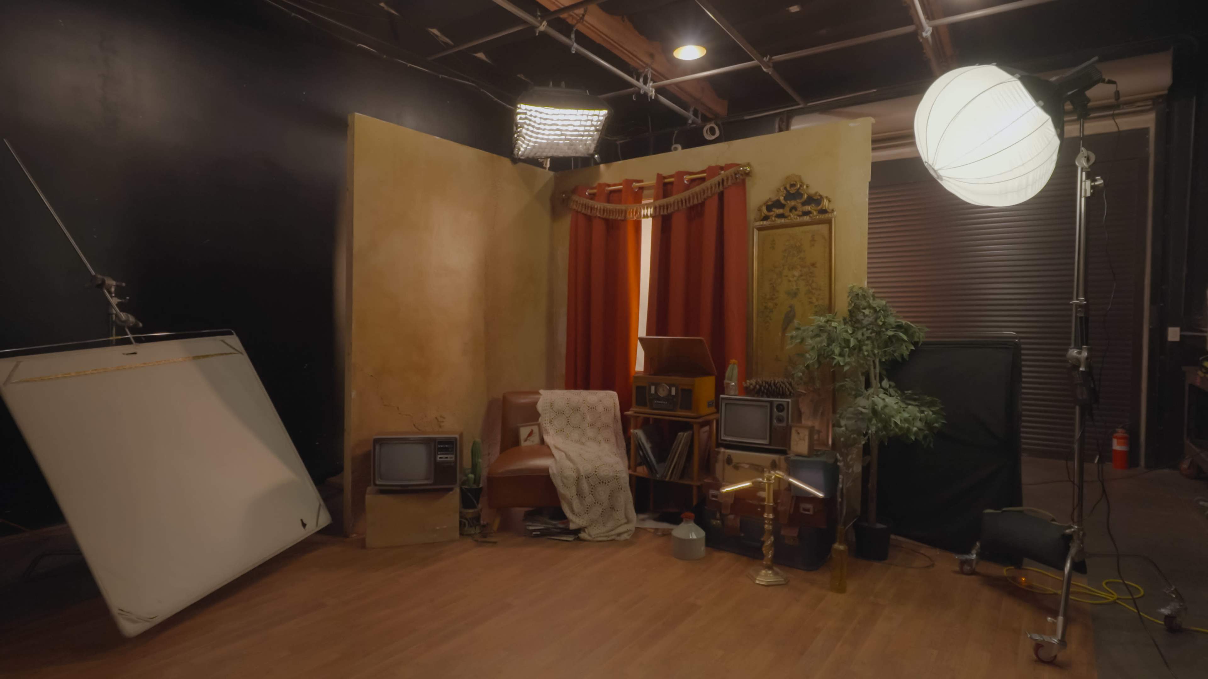 The image displays a film set with a vintage armchair, a patterned curtain, an assortment of props including old televisions, and various lighting equipment.