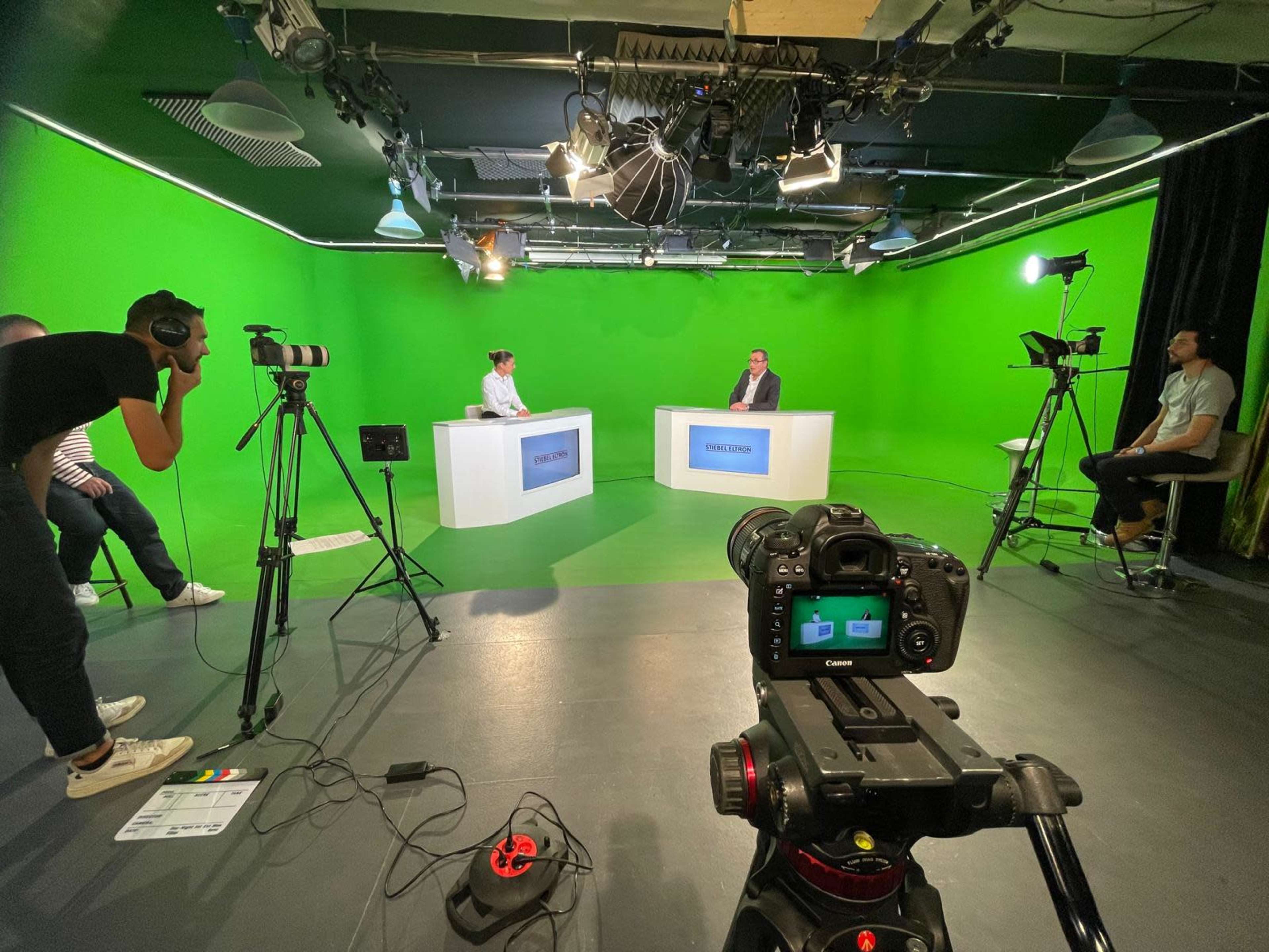 The image shows a television studio with a green screen background, where two people sit at white desks while a camera is positioned in the foreground, and additional crew members operate equipment around the set.