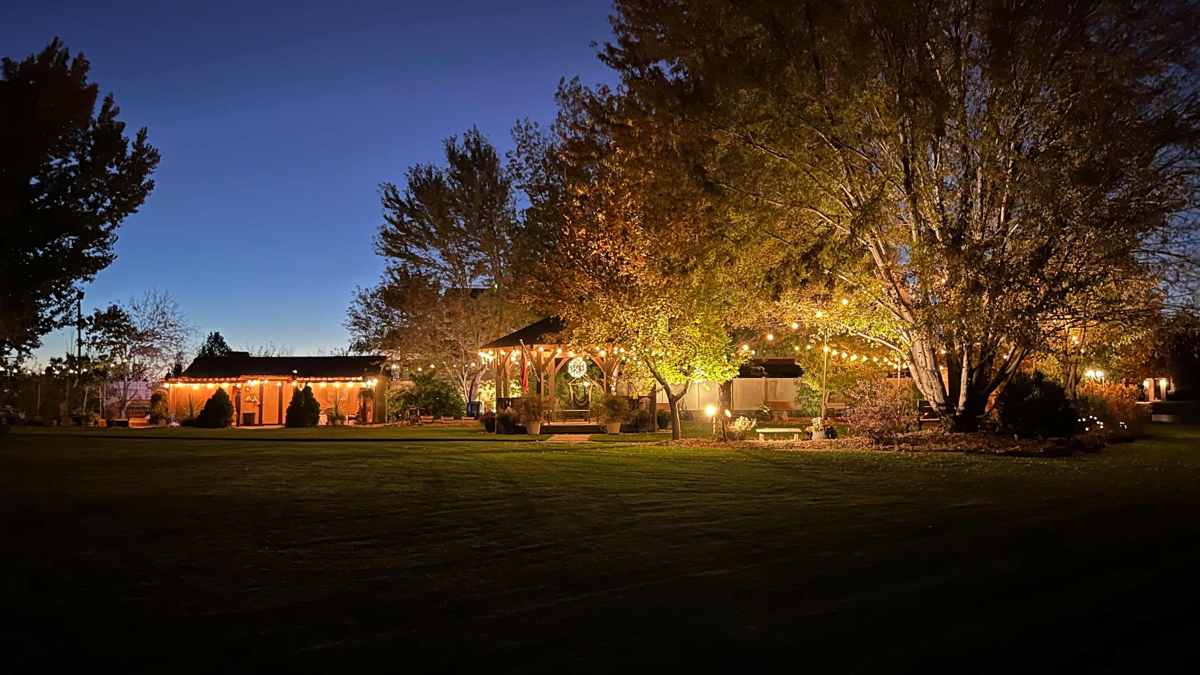 A well-lit outdoor setting at dusk features a landscaped area with trees and a gazebo surrounded by various light decorations.