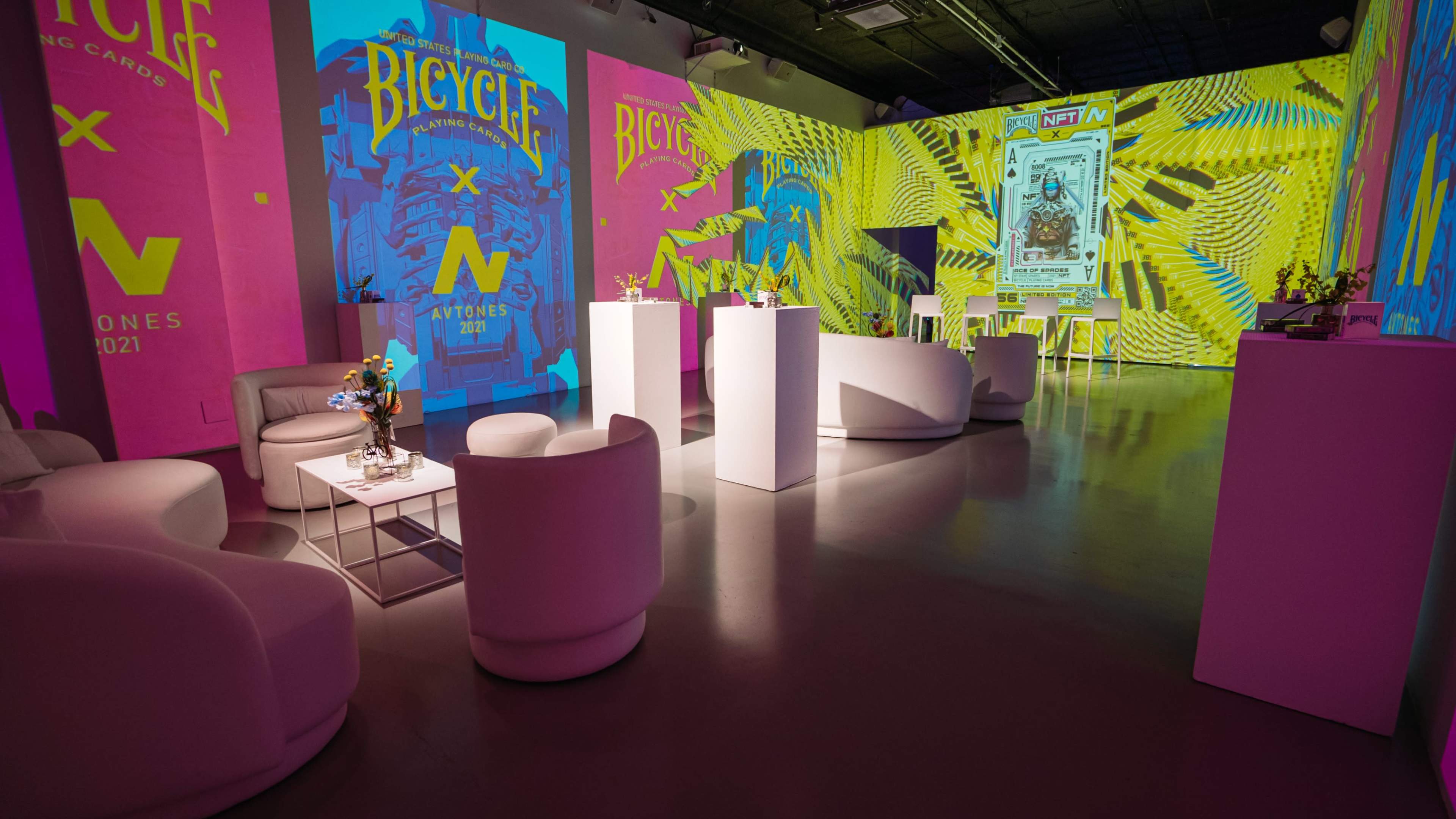 A modern event space with white furniture and vibrant, colorful projections on the walls featuring designs related to bicycles and NFTs.