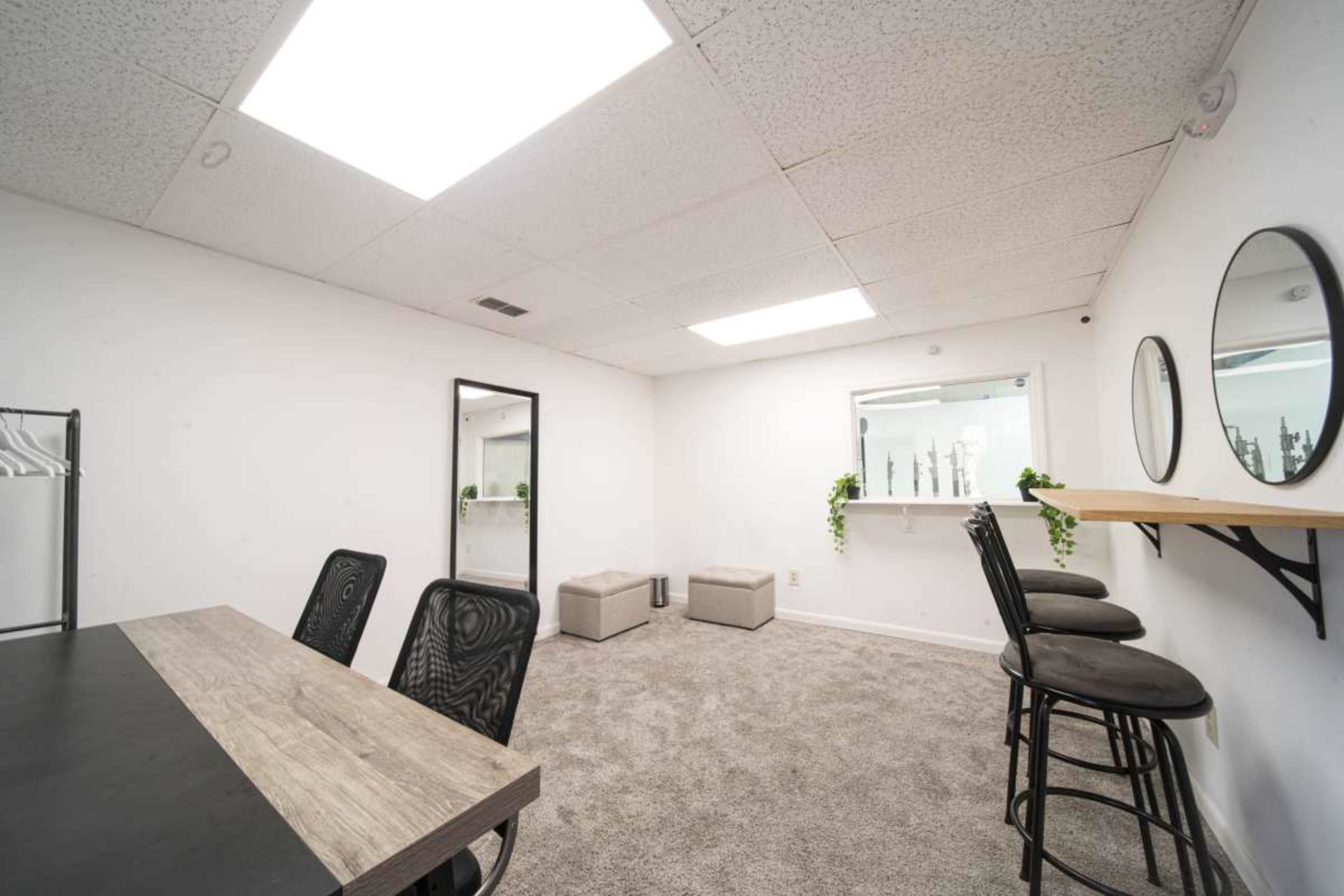 The image shows a minimalist office space with a gray carpet, a wooden table, three bar stools, and mirrors mounted on the walls.