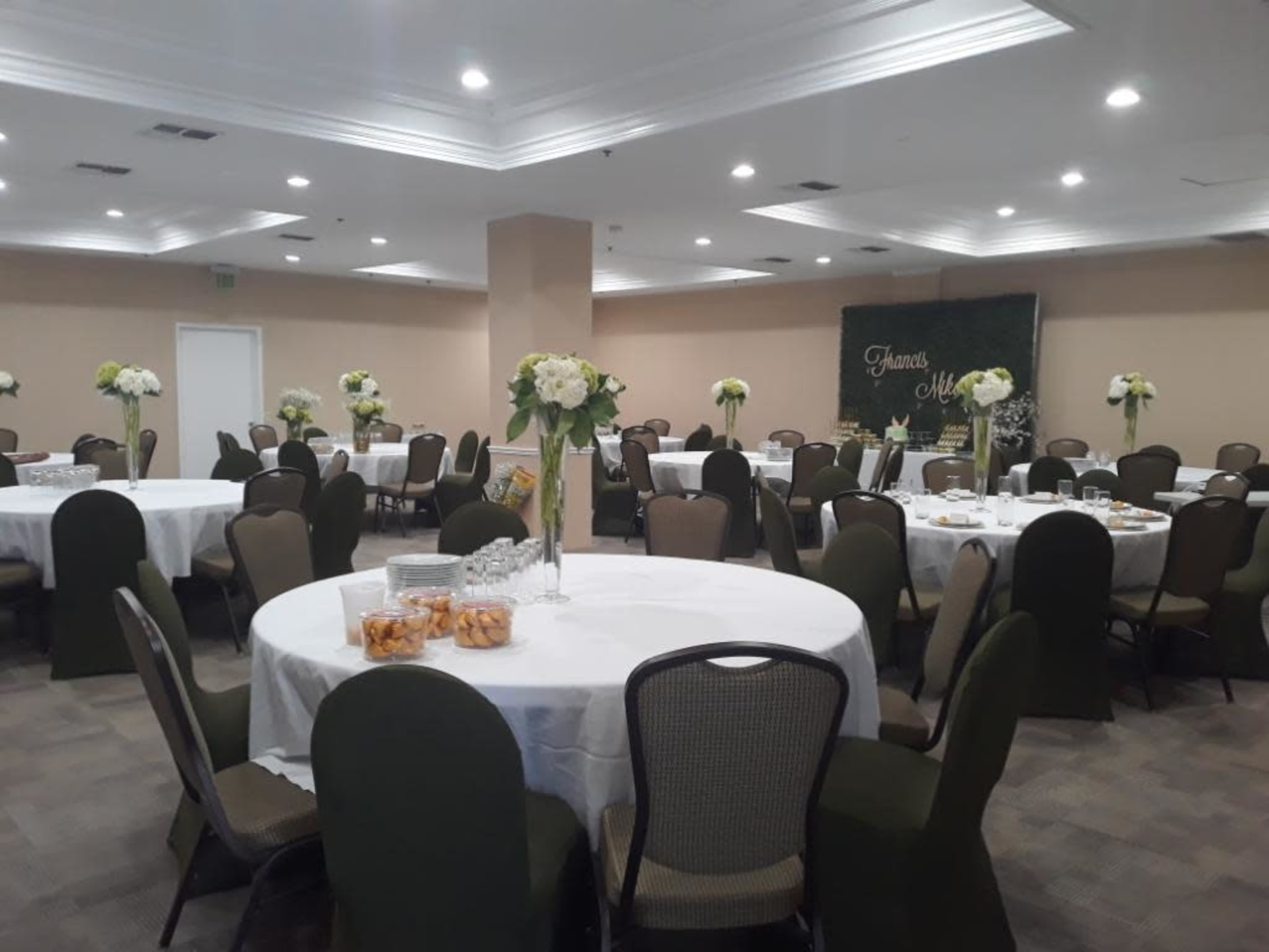 A banquet room is set up with round tables covered in white tablecloths and topped with floral centerpieces, surrounded by green chairs.