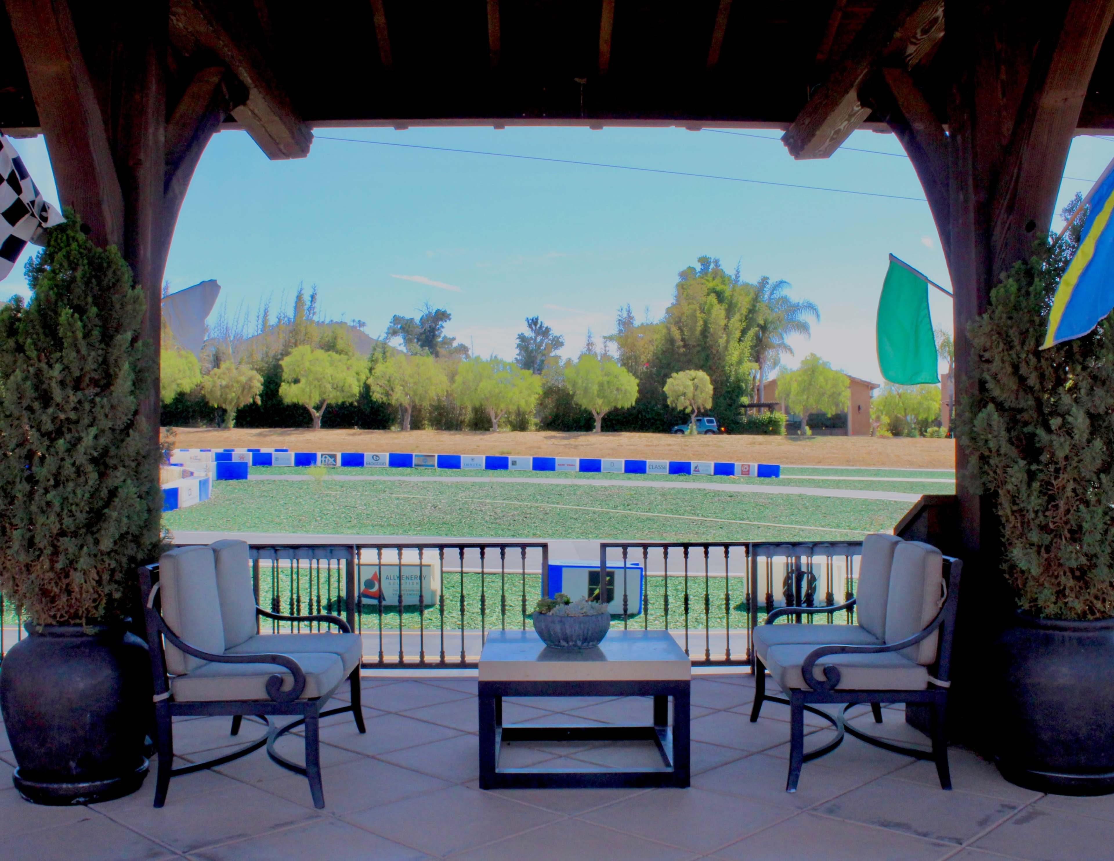 The image shows a porch with two chairs and a table overlooking a green field surrounded by blue and white barriers.
