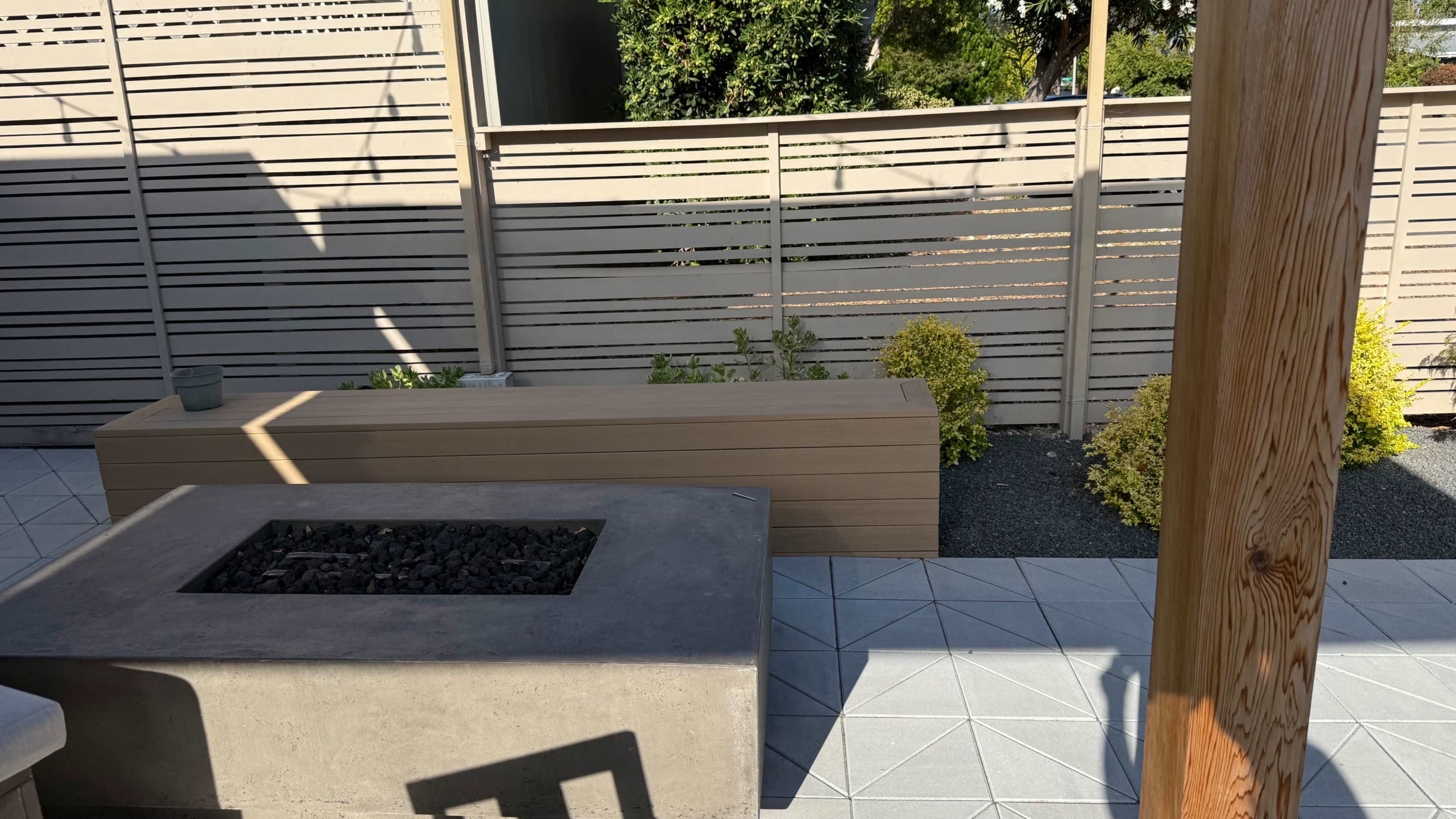 The image shows a modern outdoor space with a fire pit made of concrete, a wooden bench, and landscaping featuring small shrubs behind a slatted fence.
