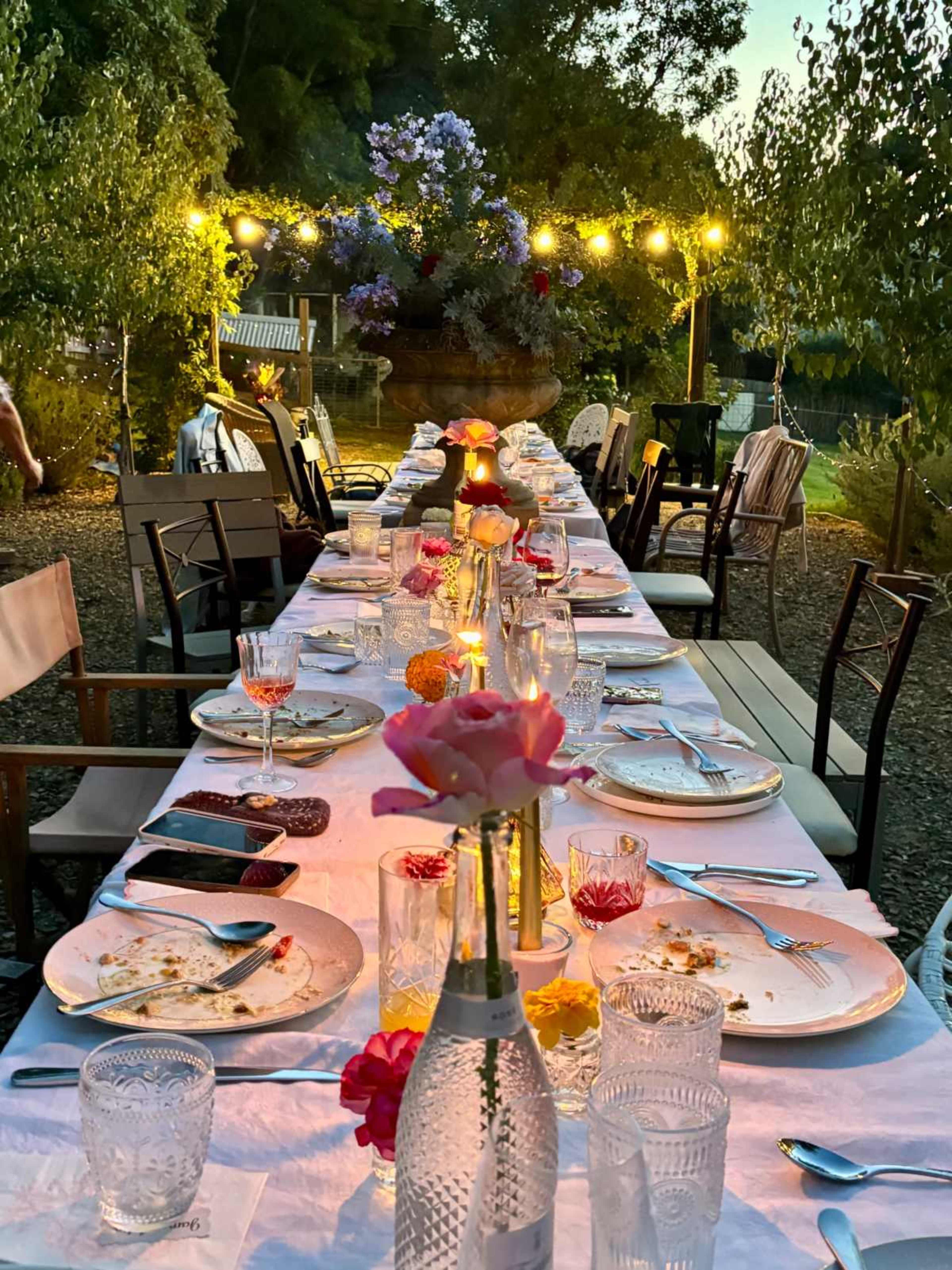 A long dining table is set outside with elegant tableware and floral arrangements, illuminated by string lights and surrounded by greenery.
