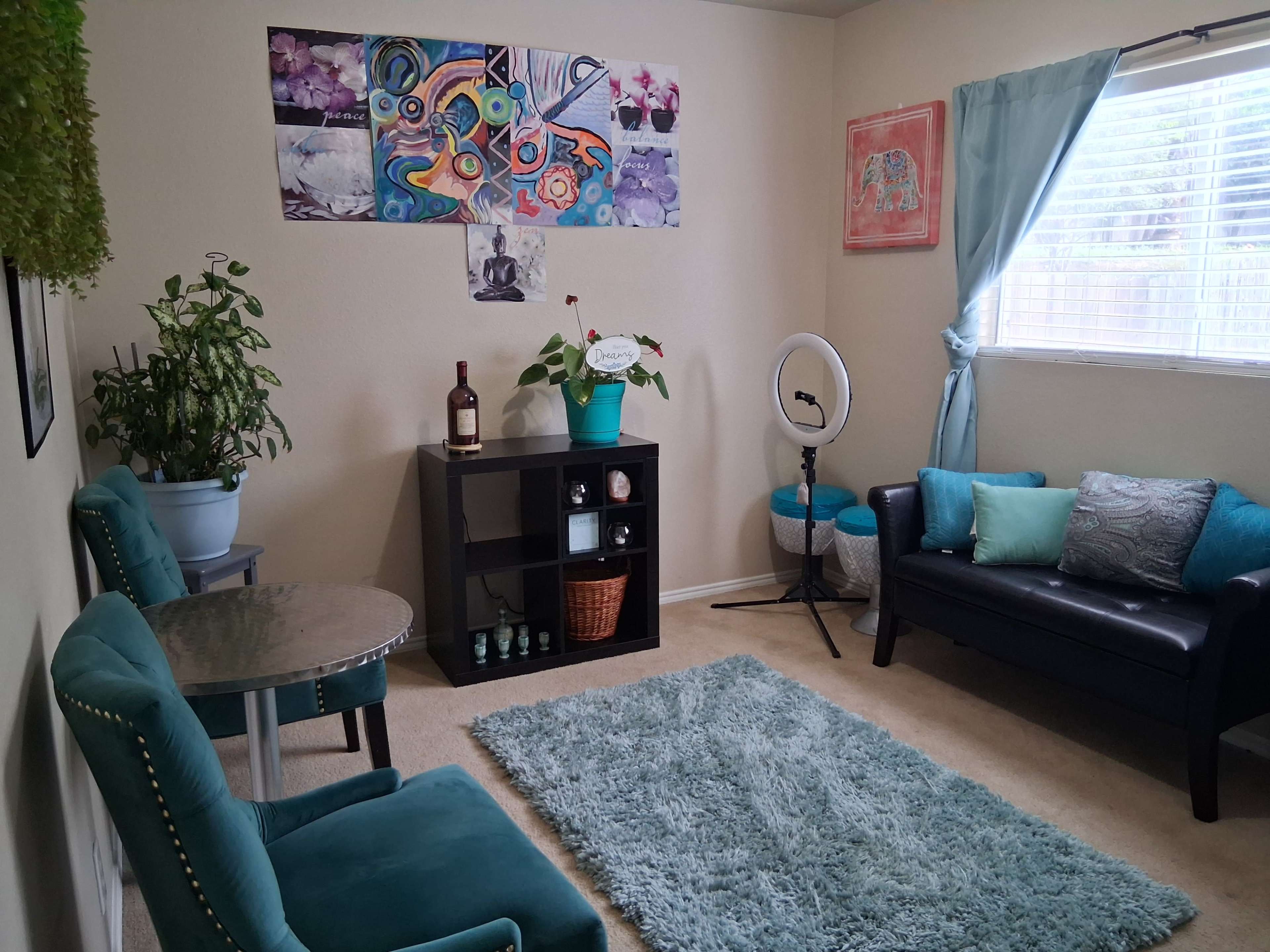 The image shows a cozy living room with teal furniture, a small round table, a plant, and vibrant wall art.