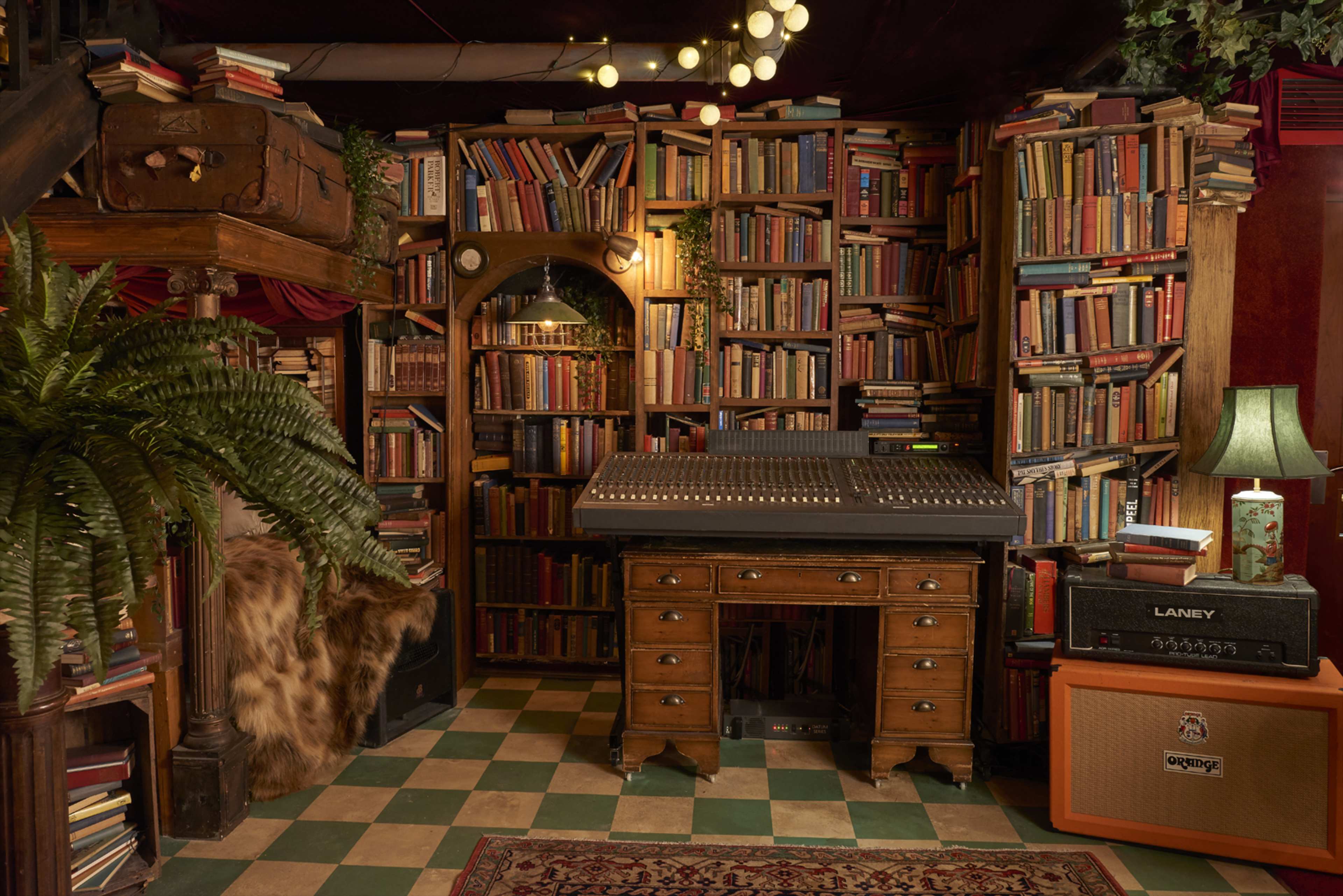 The image shows a cozy room filled with tall bookshelves packed with books, a vintage mixing console on a wooden desk, a green lamp, and a guitar amplifier beside a patterned rug.