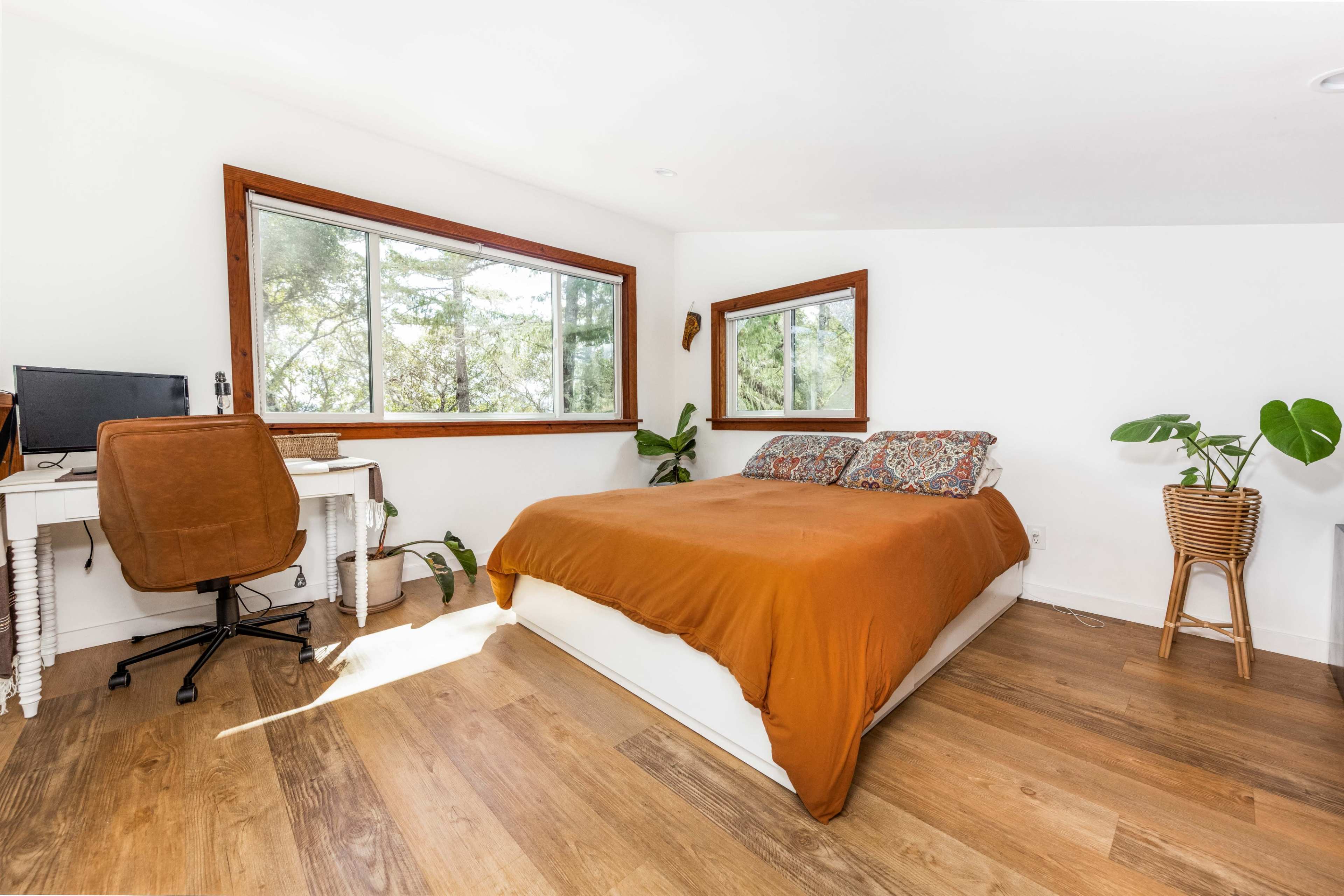 A bedroom featuring a large bed with an orange blanket, a desk with a computer, and two windows that provide natural light.