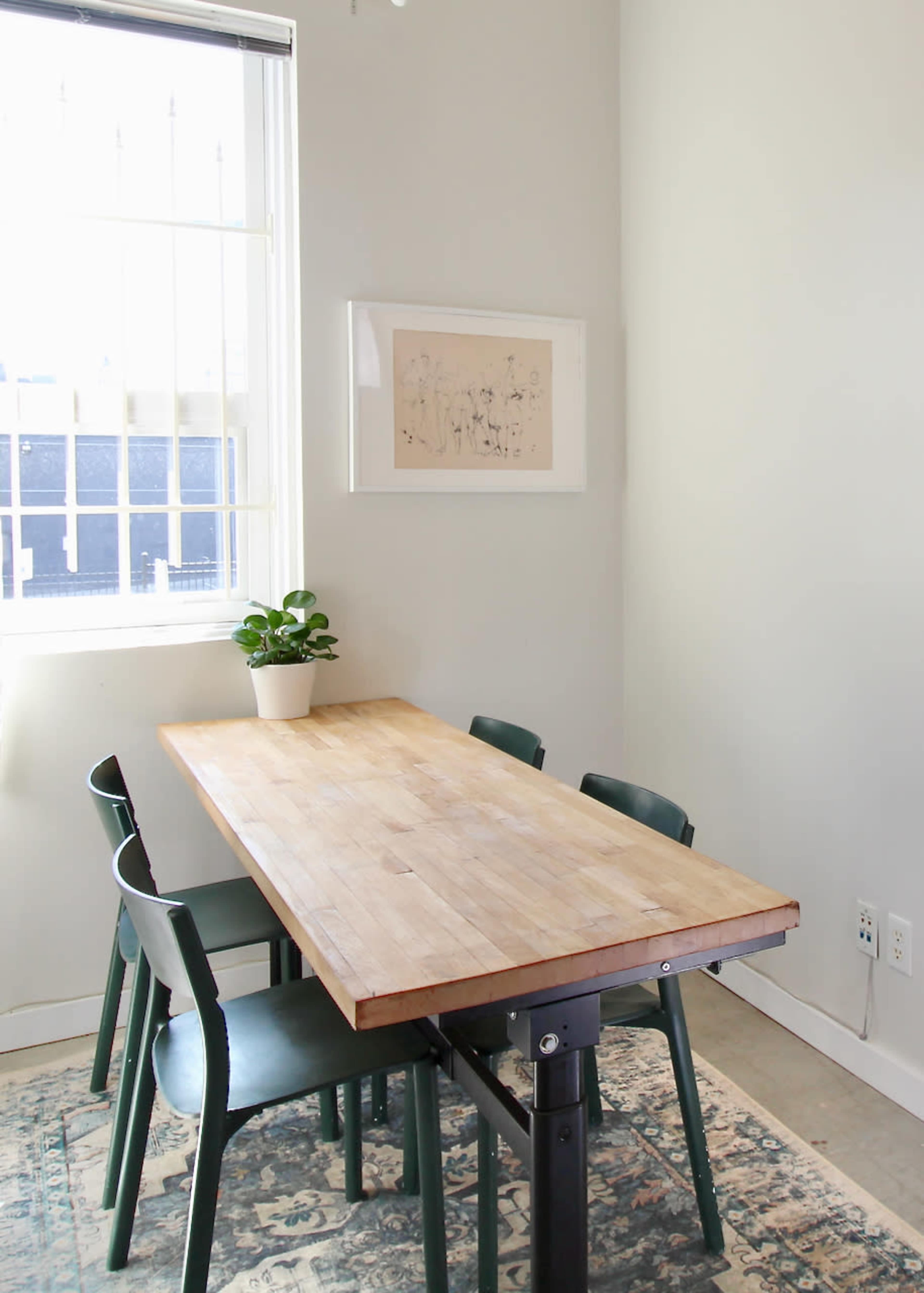 A wooden dining table with four green chairs is positioned in a corner of a room with light-colored walls and a window, featuring a small potted plant and artwork on the wall.