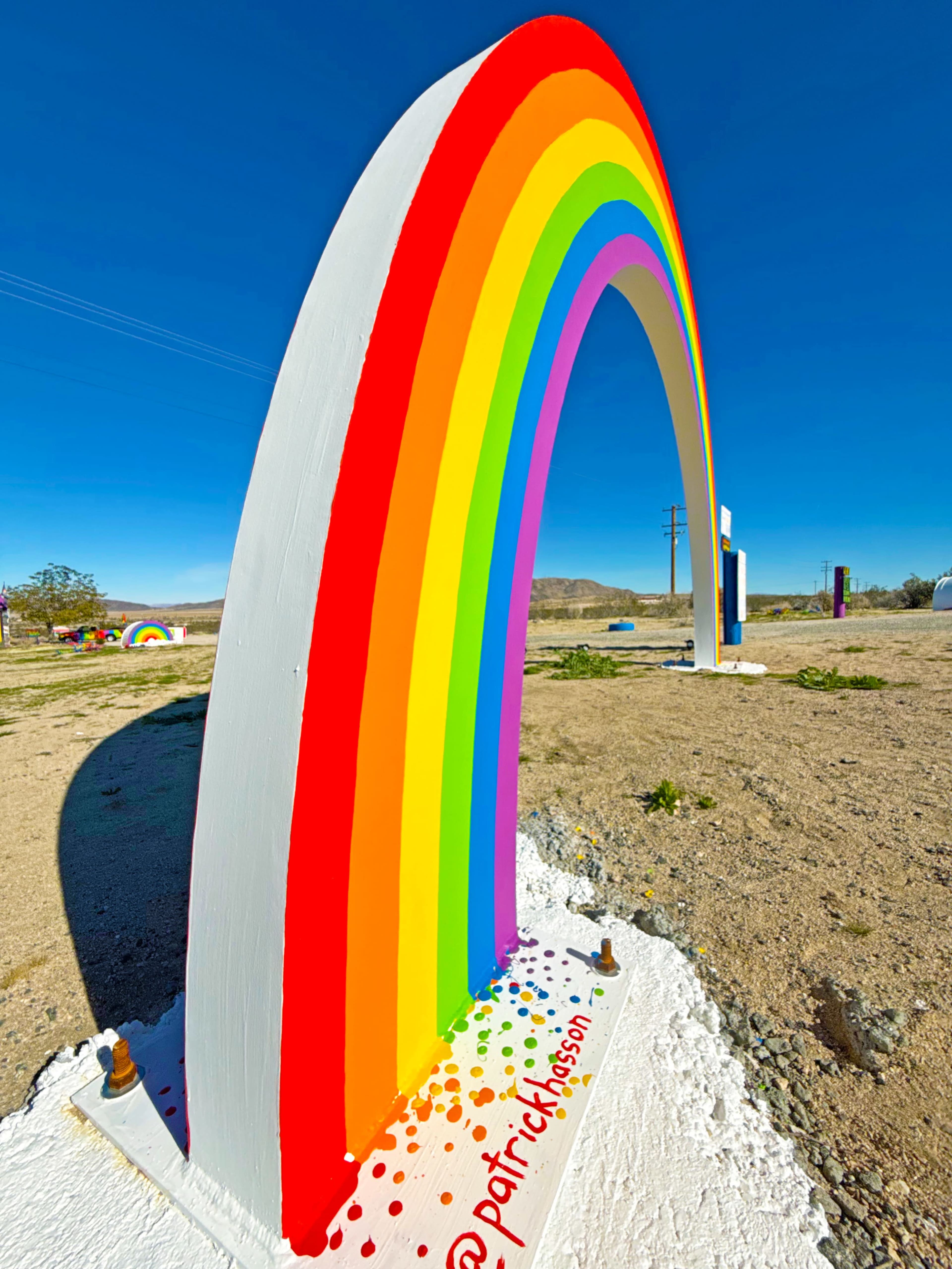 A large, colorful rainbow arch is displayed in a desert setting under a clear blue sky.