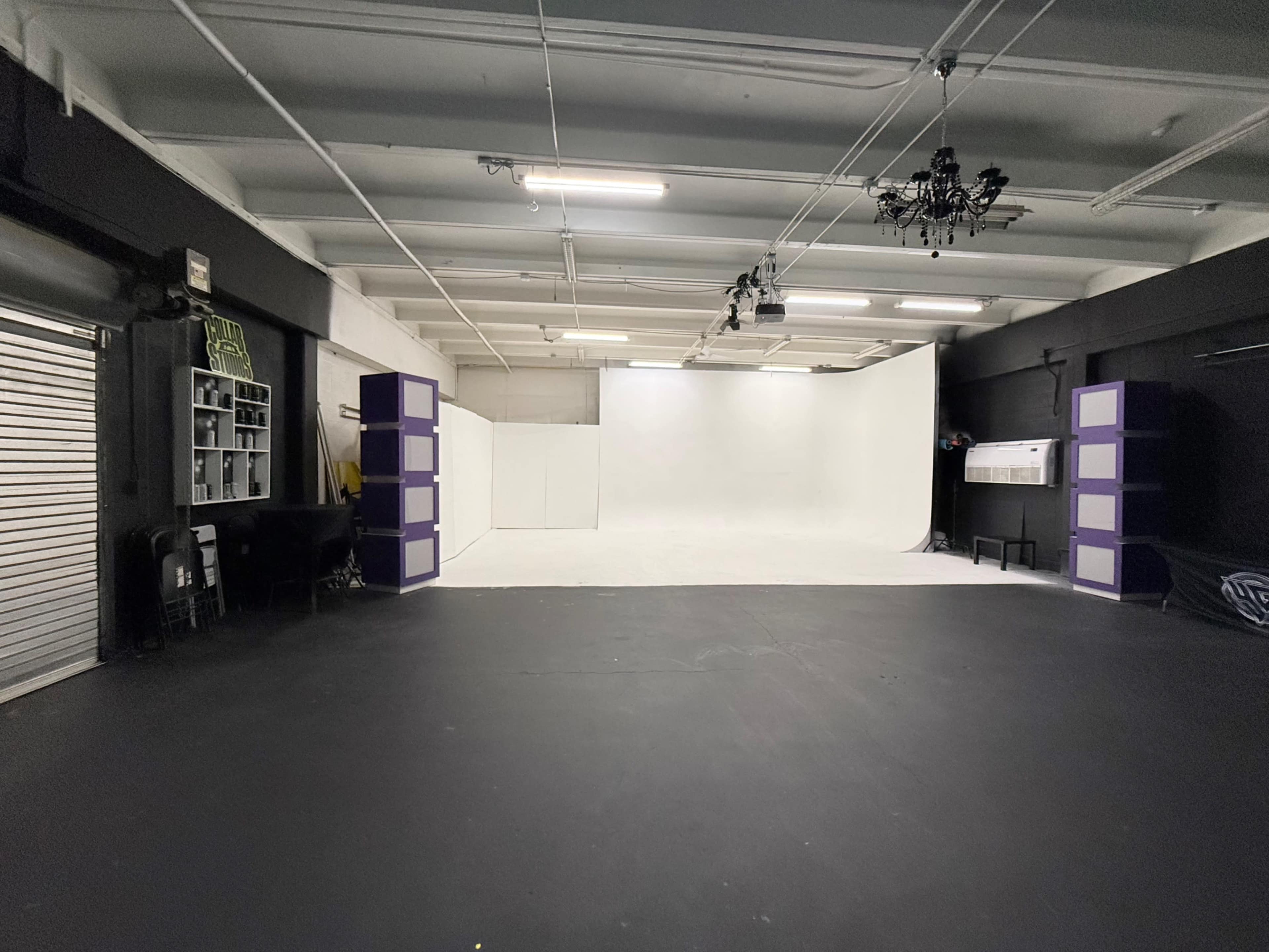 The image shows a spacious photography studio featuring a white backdrop, black flooring, and purple display structures on either side.