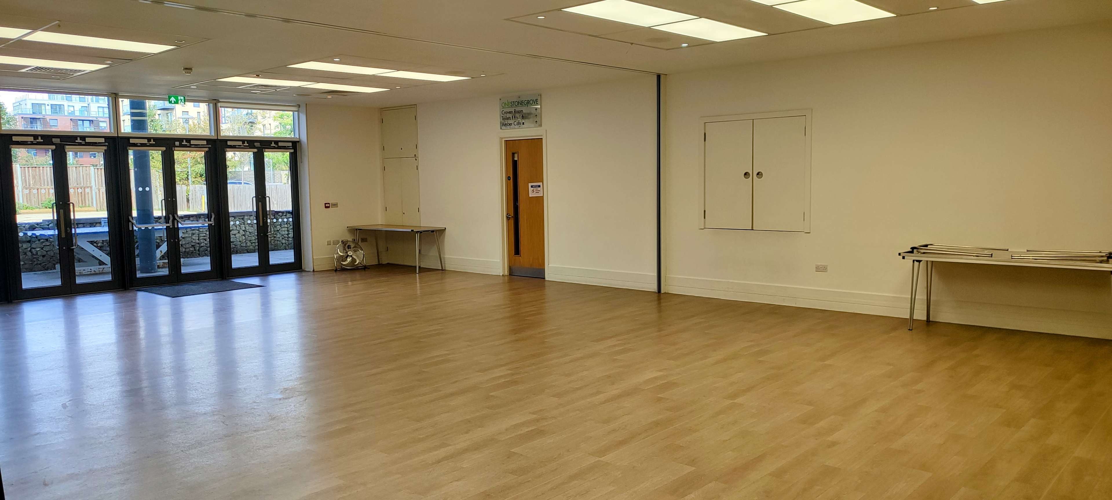 A spacious, empty room with large windows and a wooden floor, featuring two metal tables along the sides.