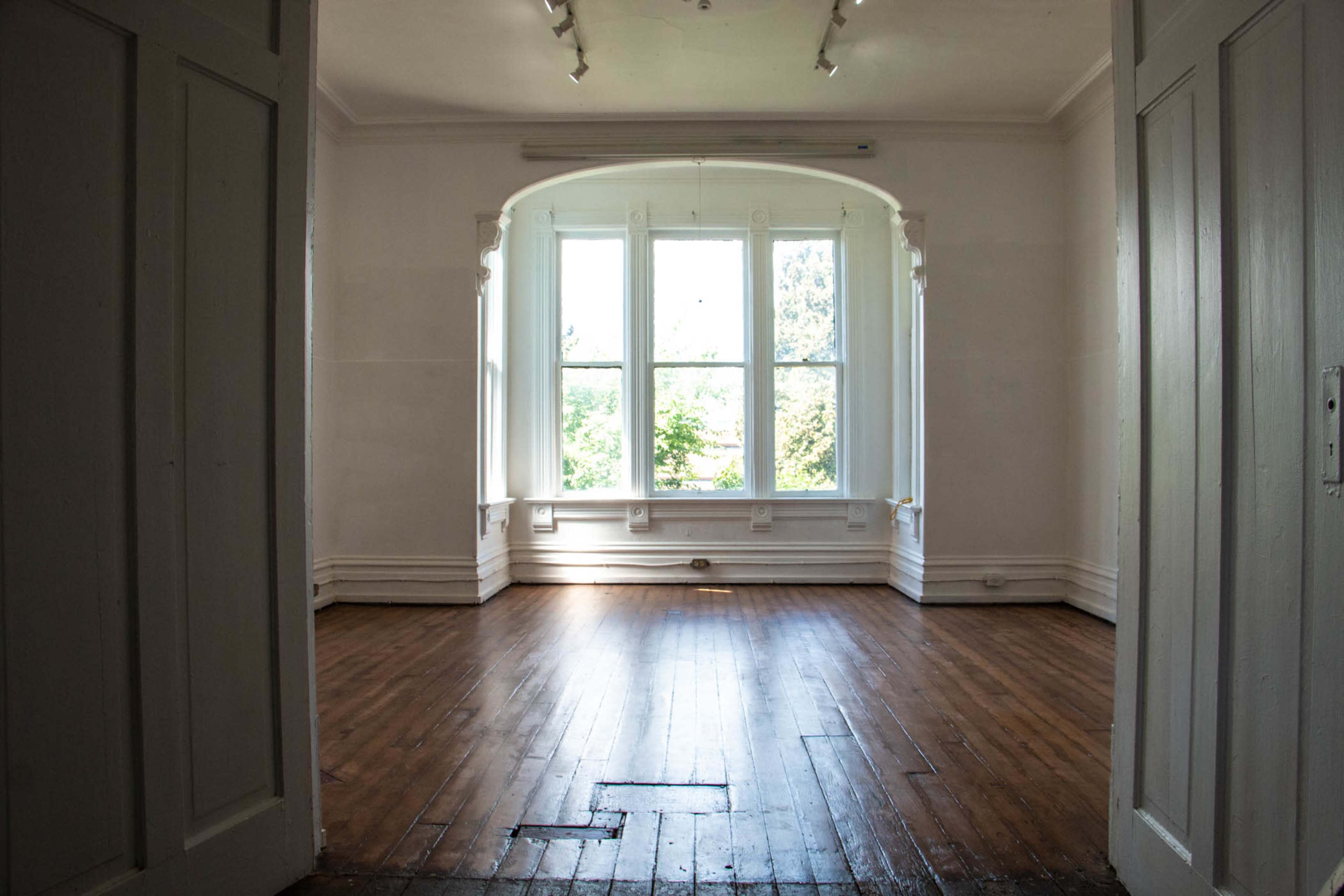 The image shows a spacious, well-lit room with wooden floors and large windows facing a green outside view.