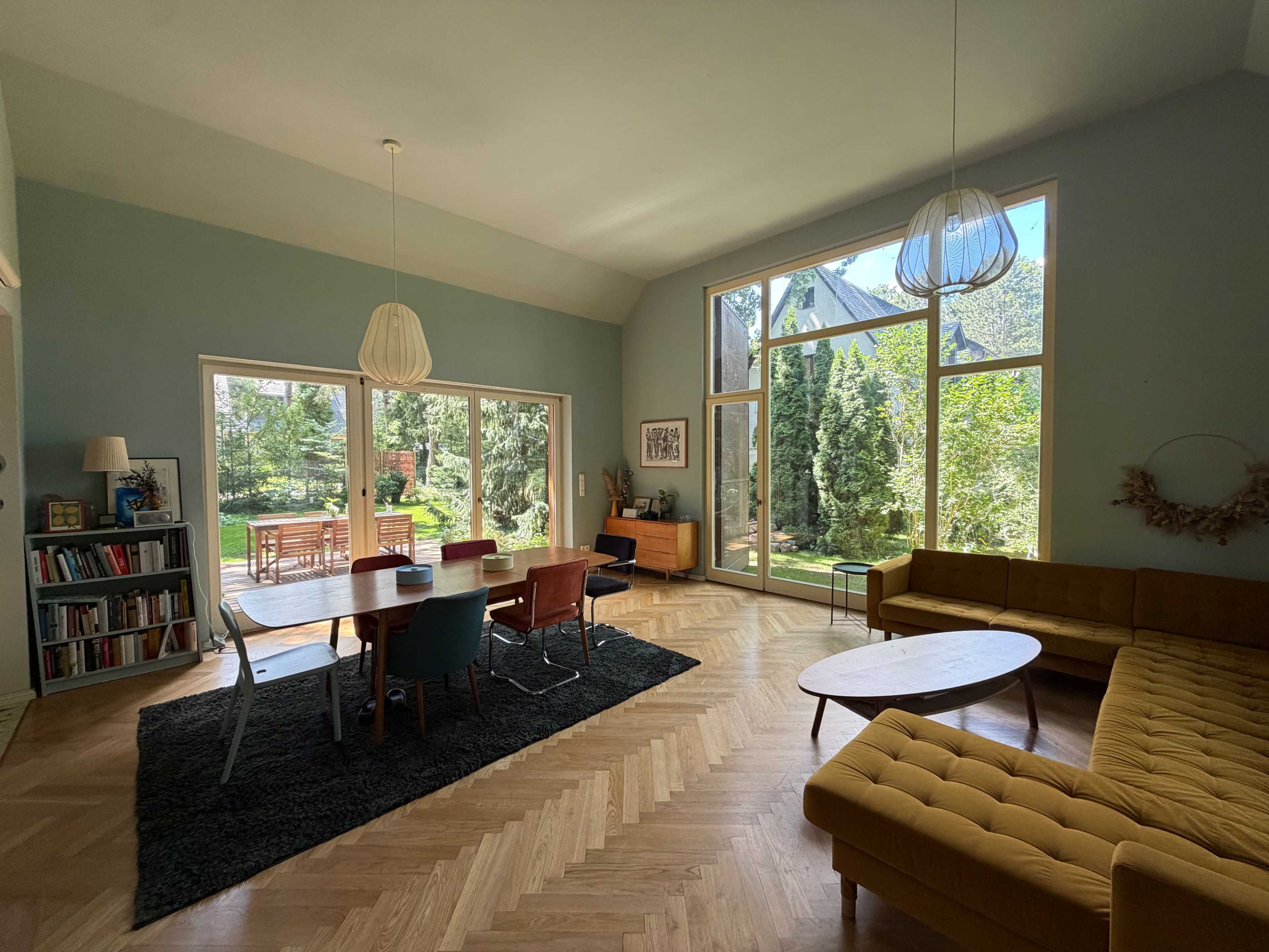 A spacious living area features a large dining table surrounded by chairs, a sectional sofa, and large windows that provide a view of a garden.