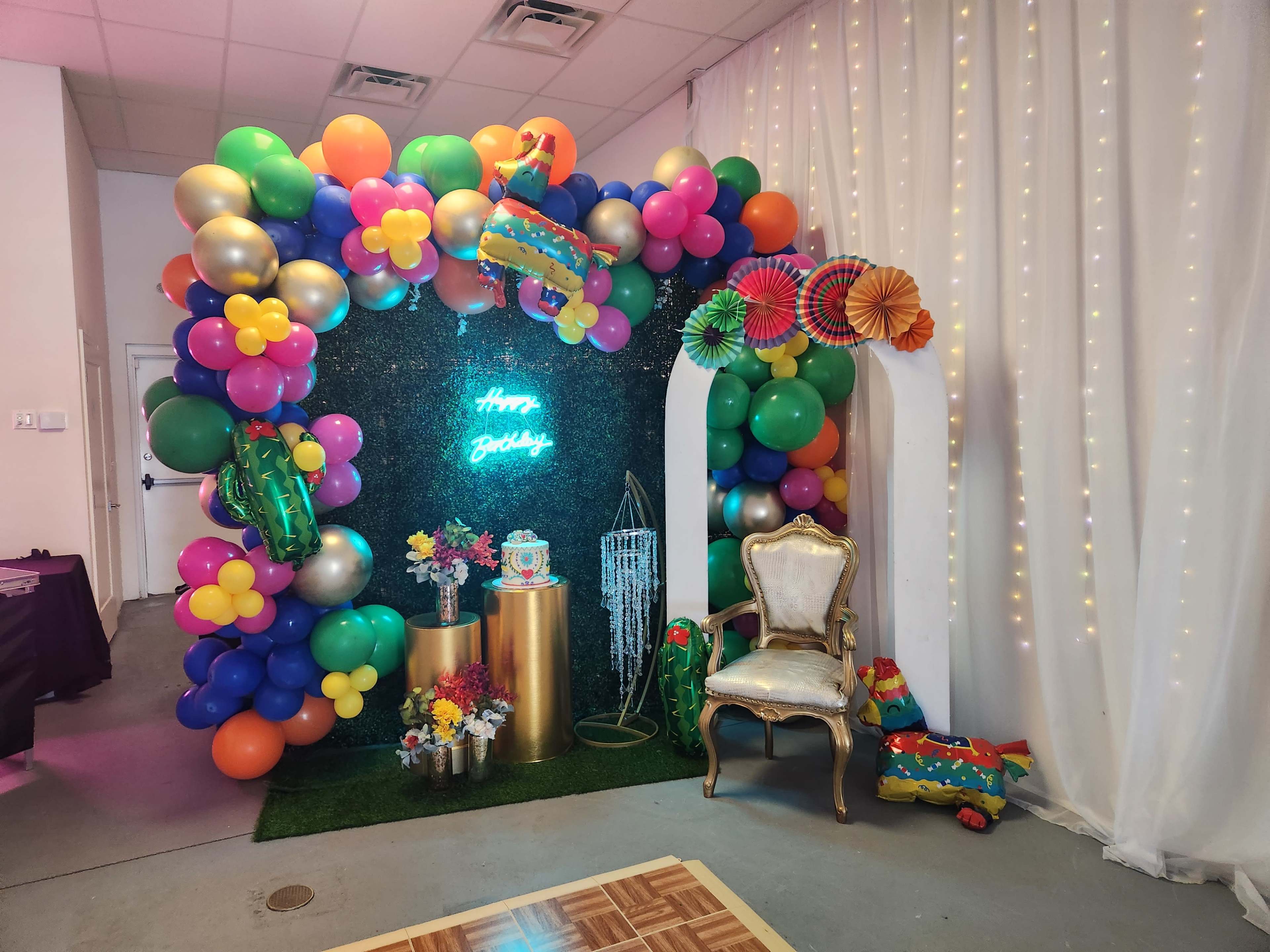 A festive birthday backdrop featuring colorful balloons, a decorated table with flowers, and a chair surrounded by party decorations.