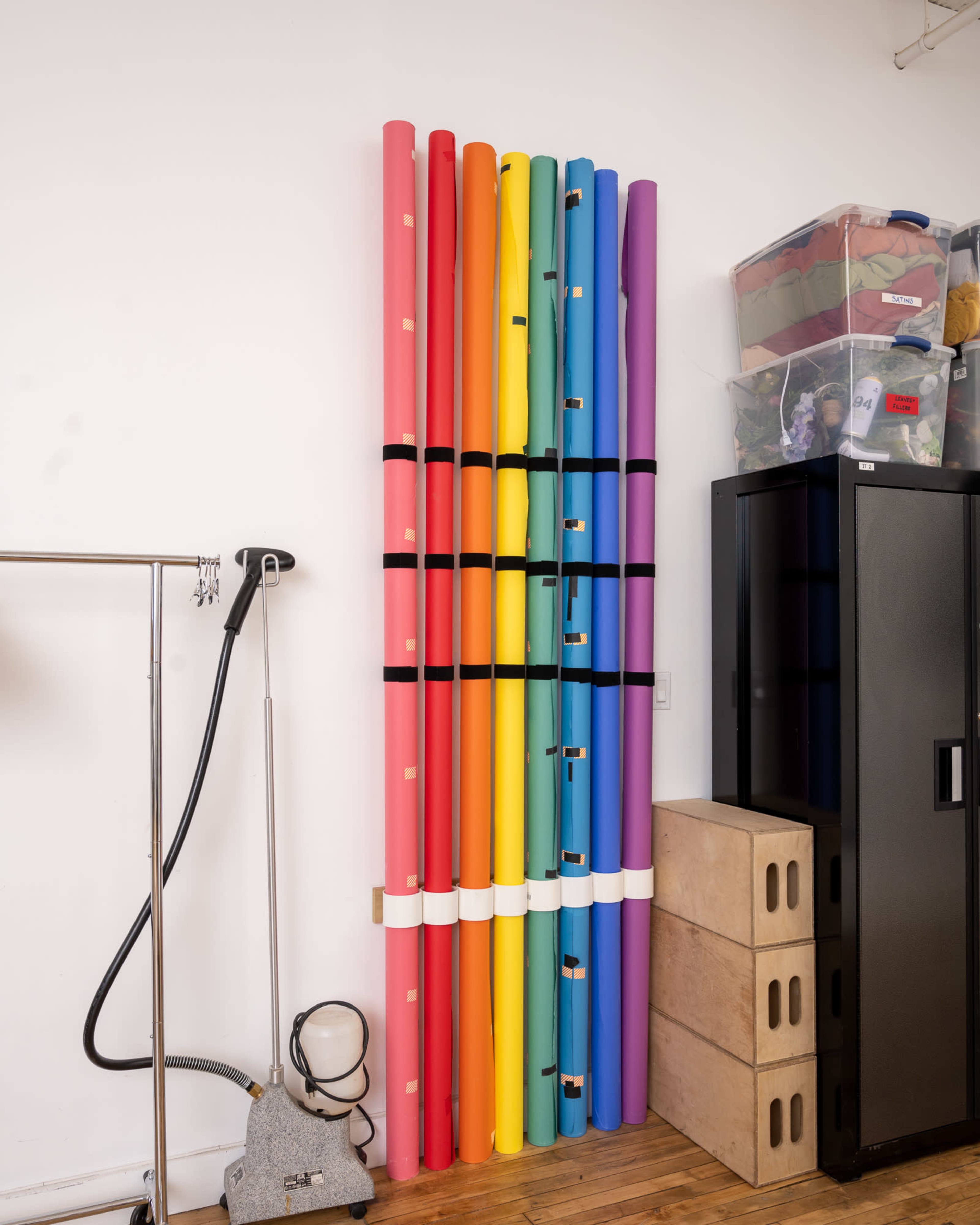 The image shows a collection of rolled background papers in various colors stored upright against a wall, next to a black cabinet and a clothing rack.