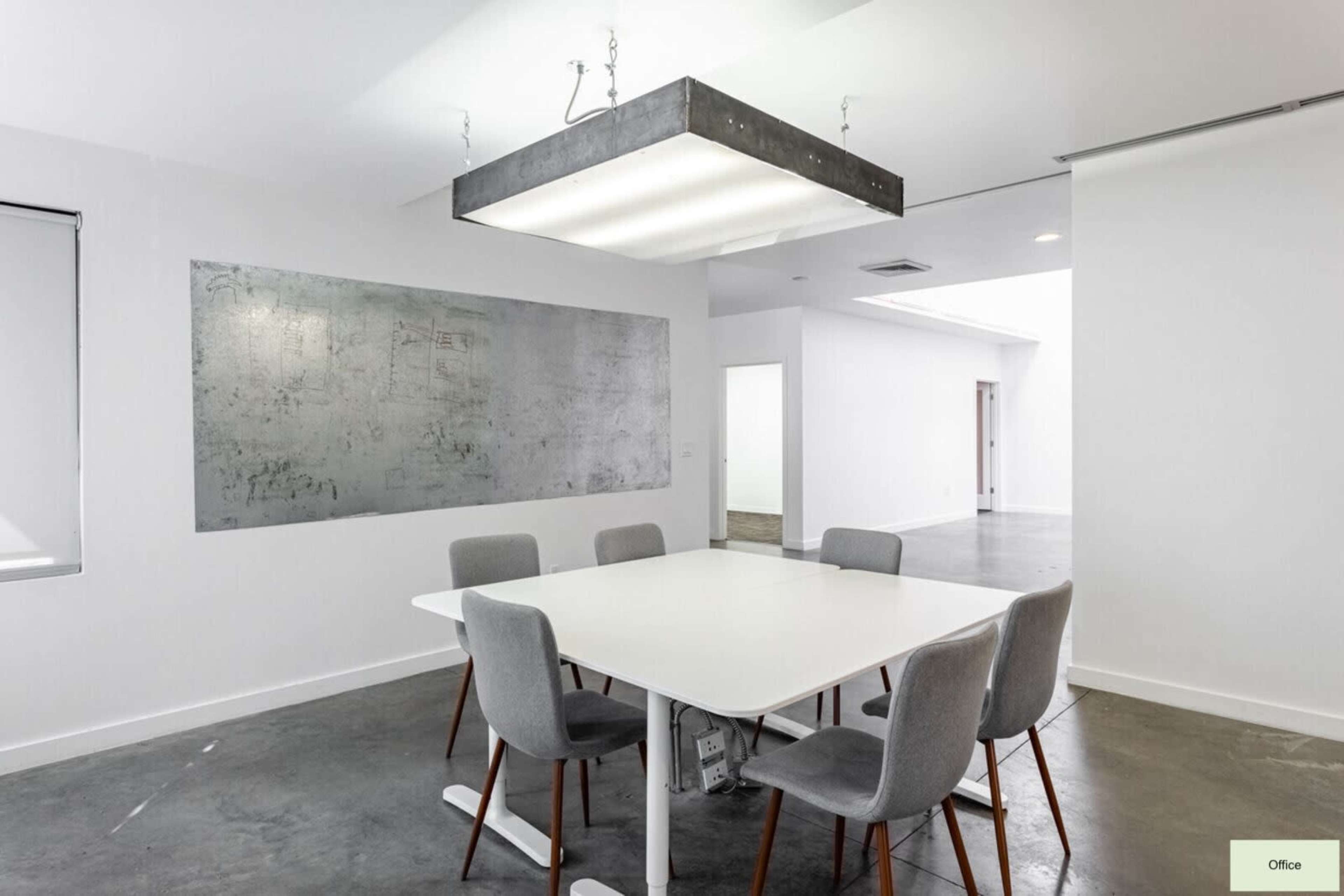 A modern office space features a white table surrounded by gray chairs, with a large light fixture hanging above it and a piece of wall art on one side.