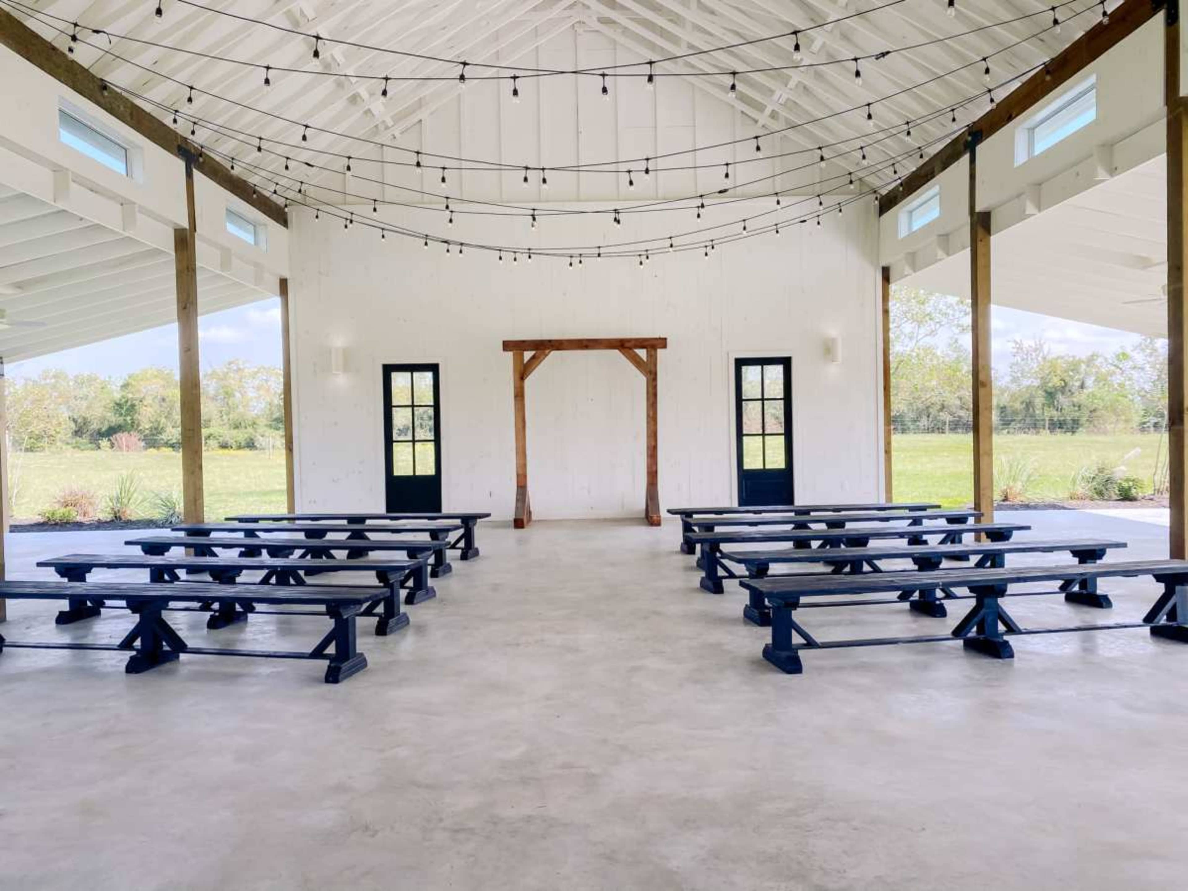 The interior of a spacious event venue features rows of wooden benches facing a wooden arch, with large windows and string lights overhead.