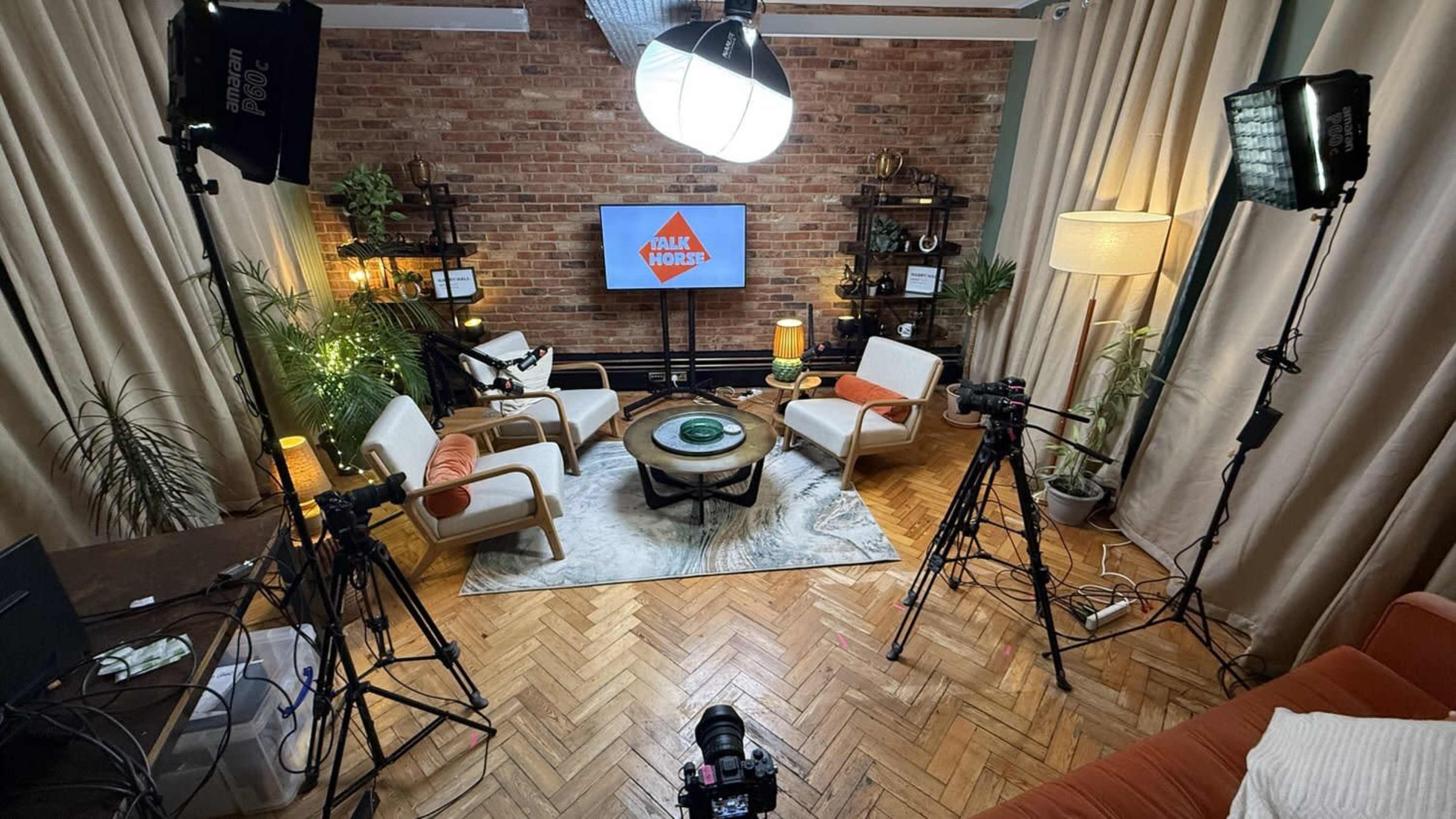 A well-lit indoor studio setup features a circular table with a green centerpiece surrounded by four chairs, flanked by plants, lighting equipment, and a television displaying the logo of "Talk Shows."