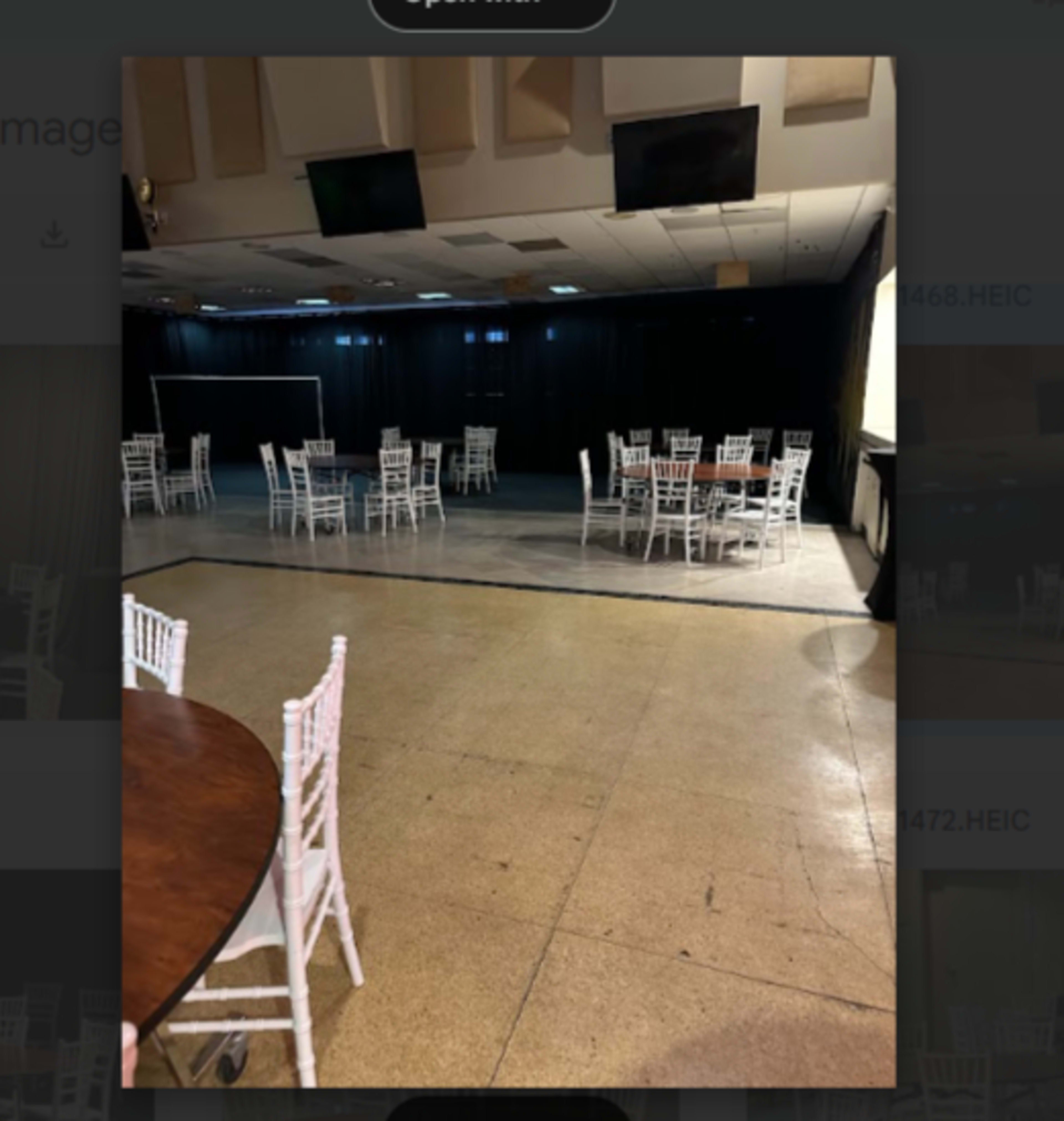The image shows a spacious event hall furnished with several round tables and white chiavari chairs, illuminated by soft overhead lighting.