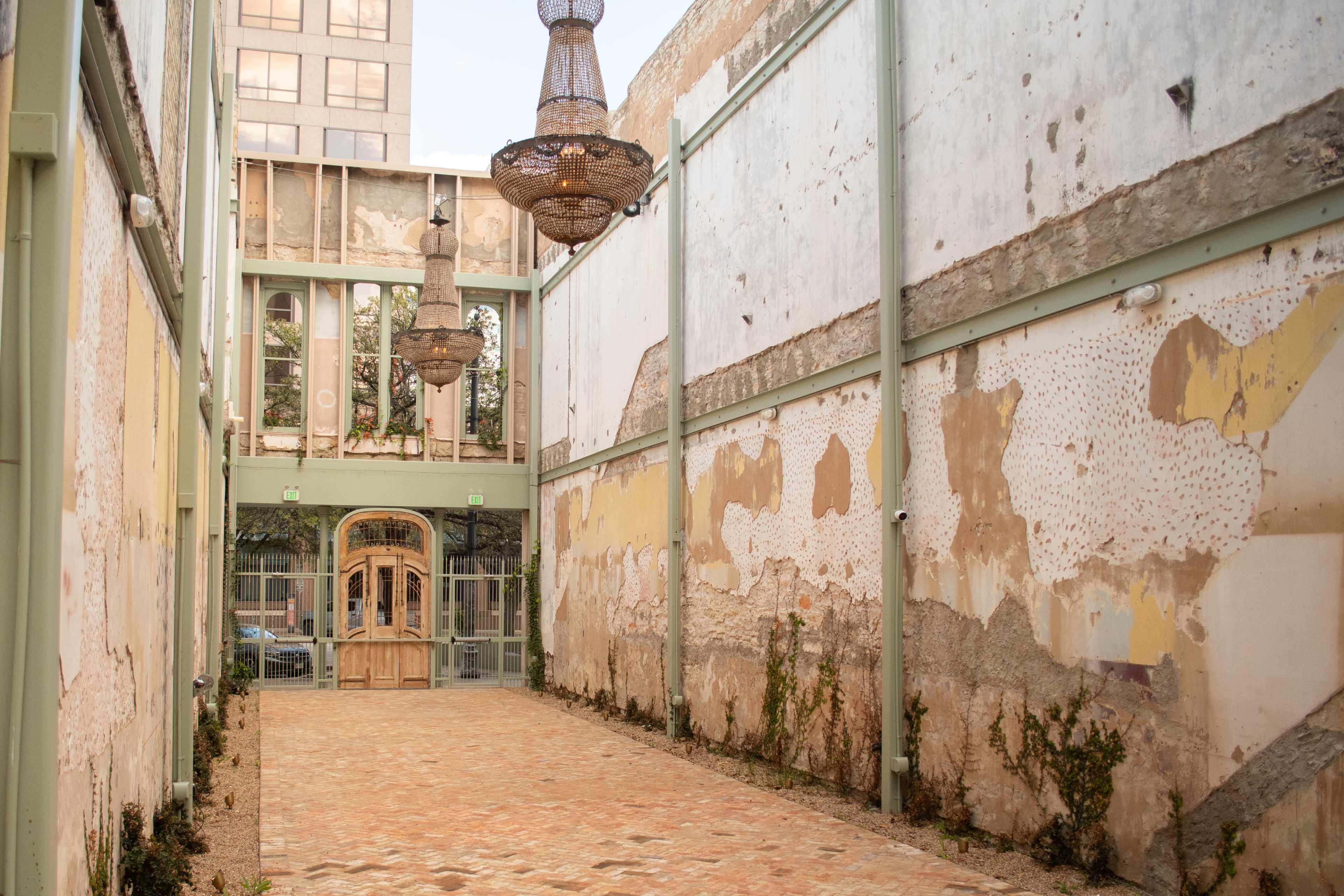 The image shows a wide, open courtyard featuring weathered walls, decorative chandeliers hanging from the ceiling, and a central door leading to an interior space.