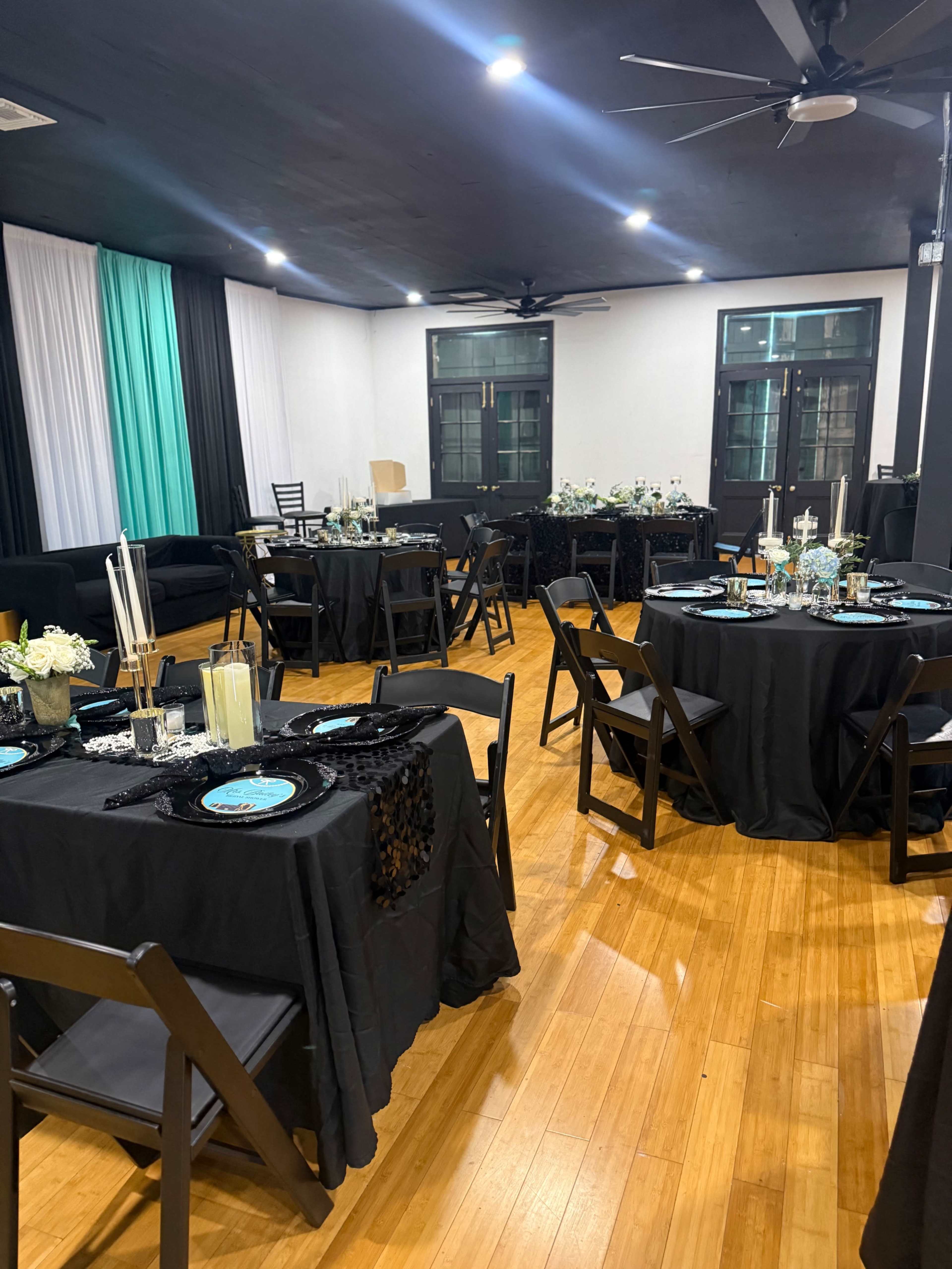 The image shows a banquet hall arranged with black tablecloths, turquoise centerpieces, and a wooden floor, featuring several tables and chairs.