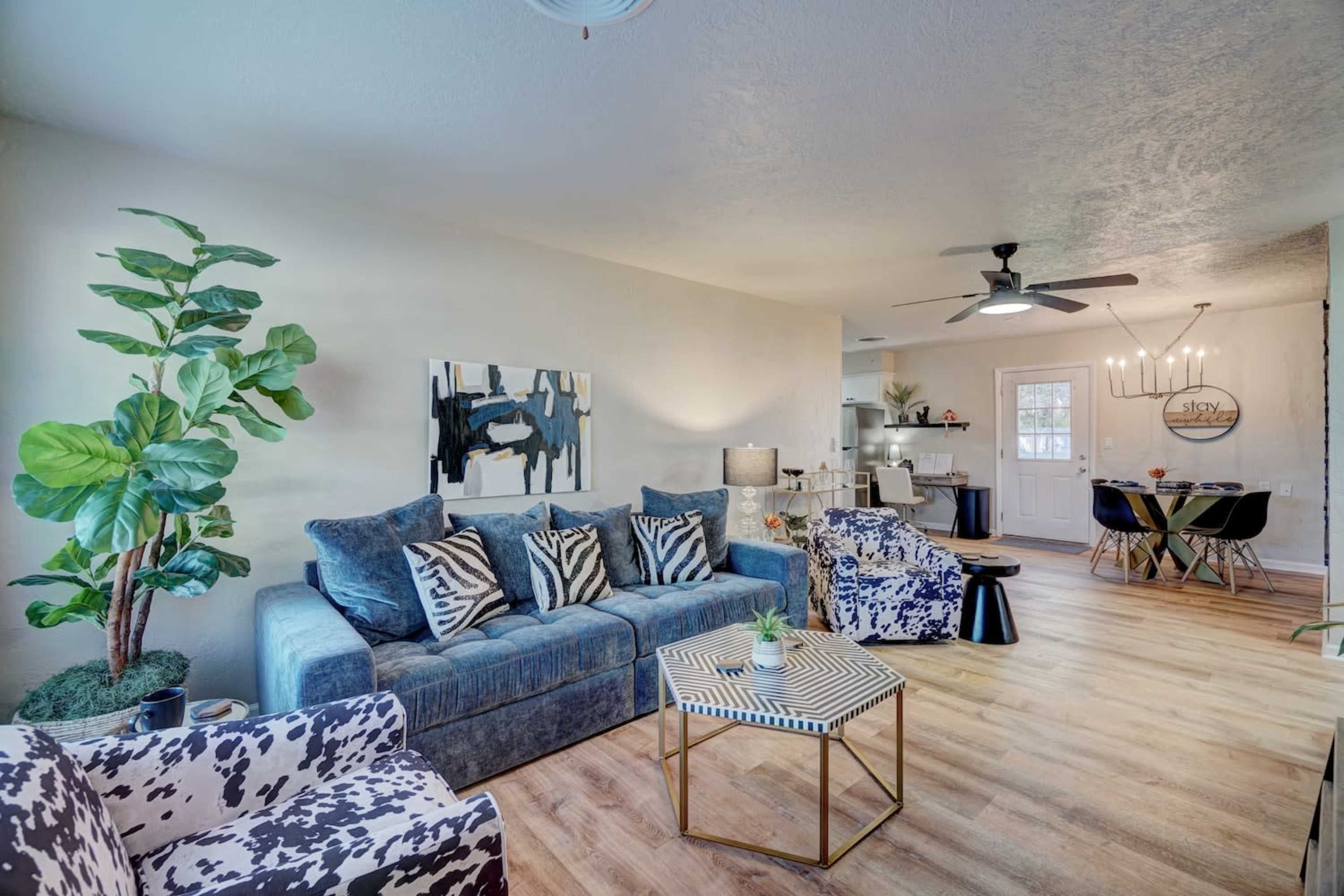 A living room features blue and patterned sofas, a coffee table, a large plant, and an open view to a dining area with a door leading outside.
