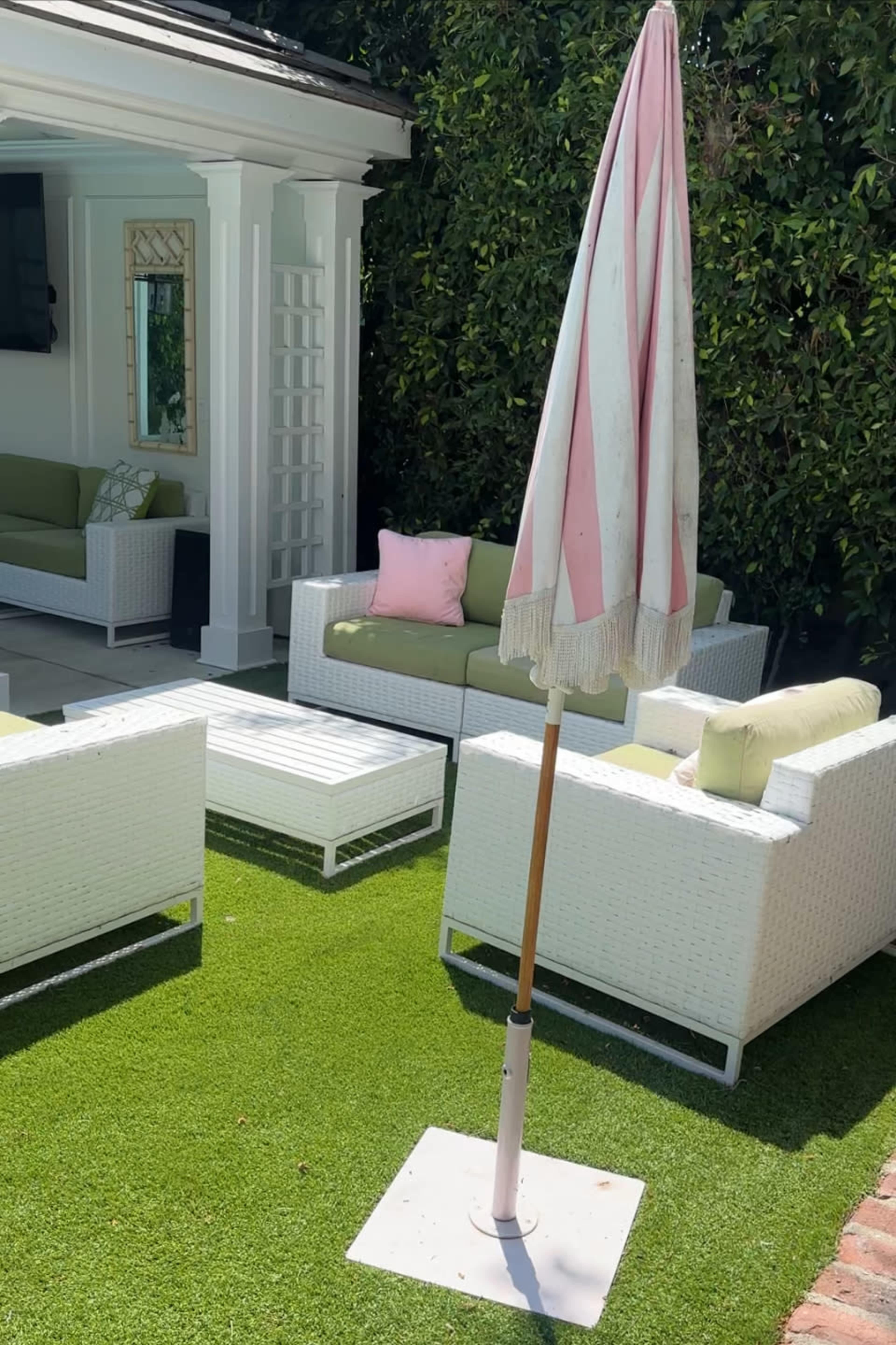 A pink and white striped umbrella stands in a base on a green lawn, beside white wicker furniture arranged in a patio setting.