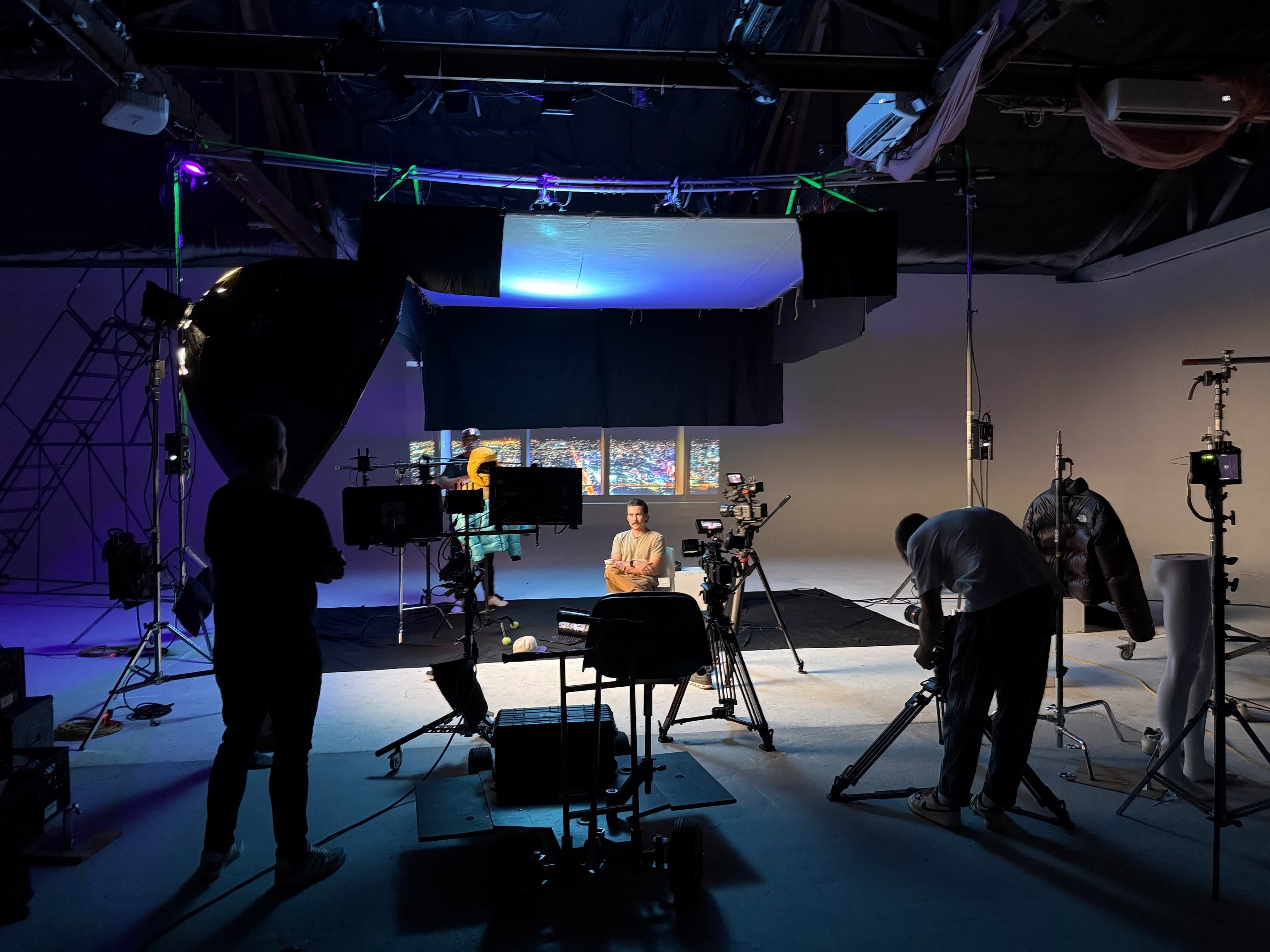The image shows a film set with various cameras, lights, and equipment, featuring a subject seated in the center.