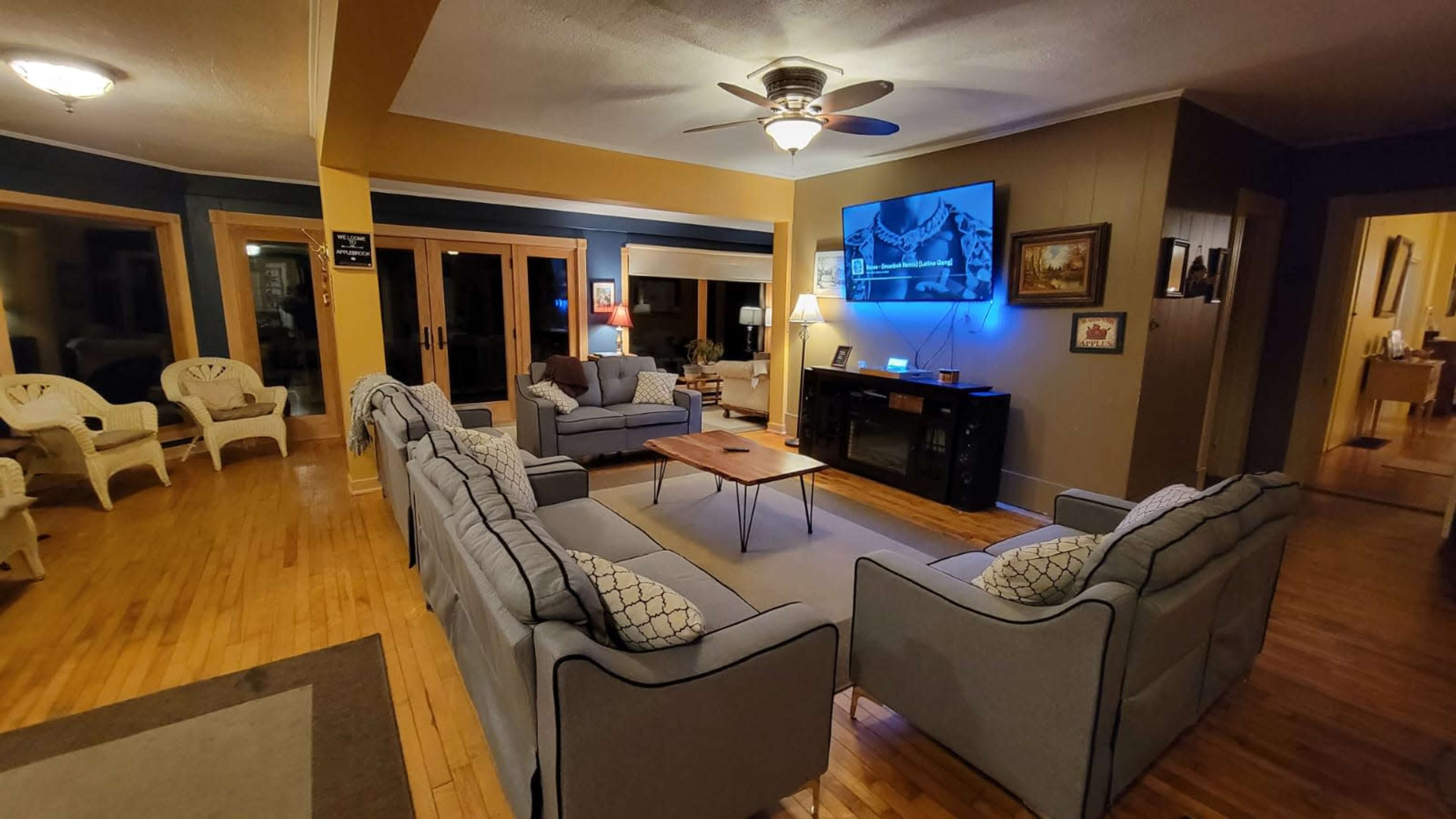 The living room features multiple seating areas, including two blue sofas and a coffee table, with a large screen mounted on the wall and sliding glass doors leading to an outdoor space.