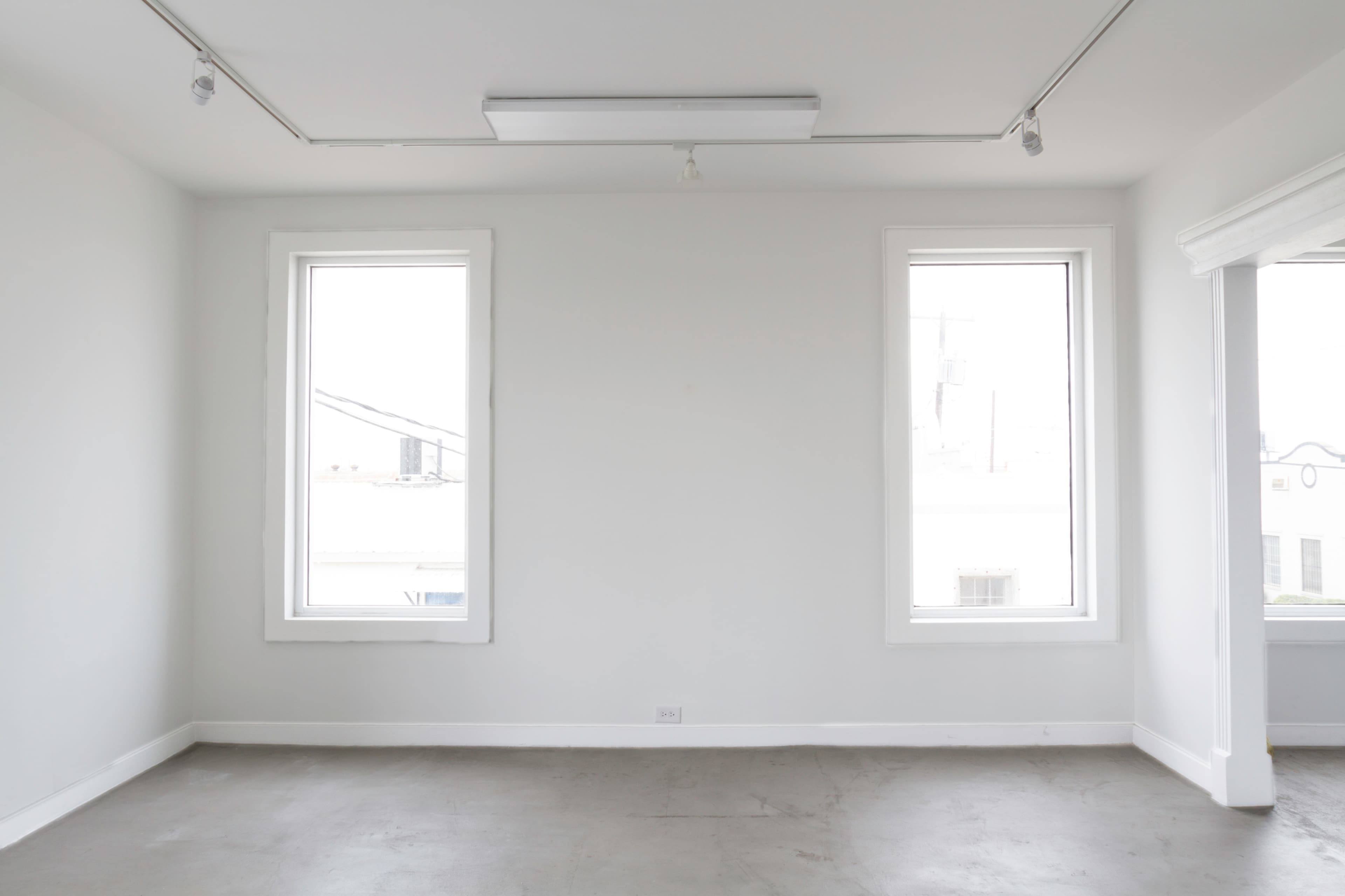 The image shows a sparsely furnished room with two windows and bare walls.