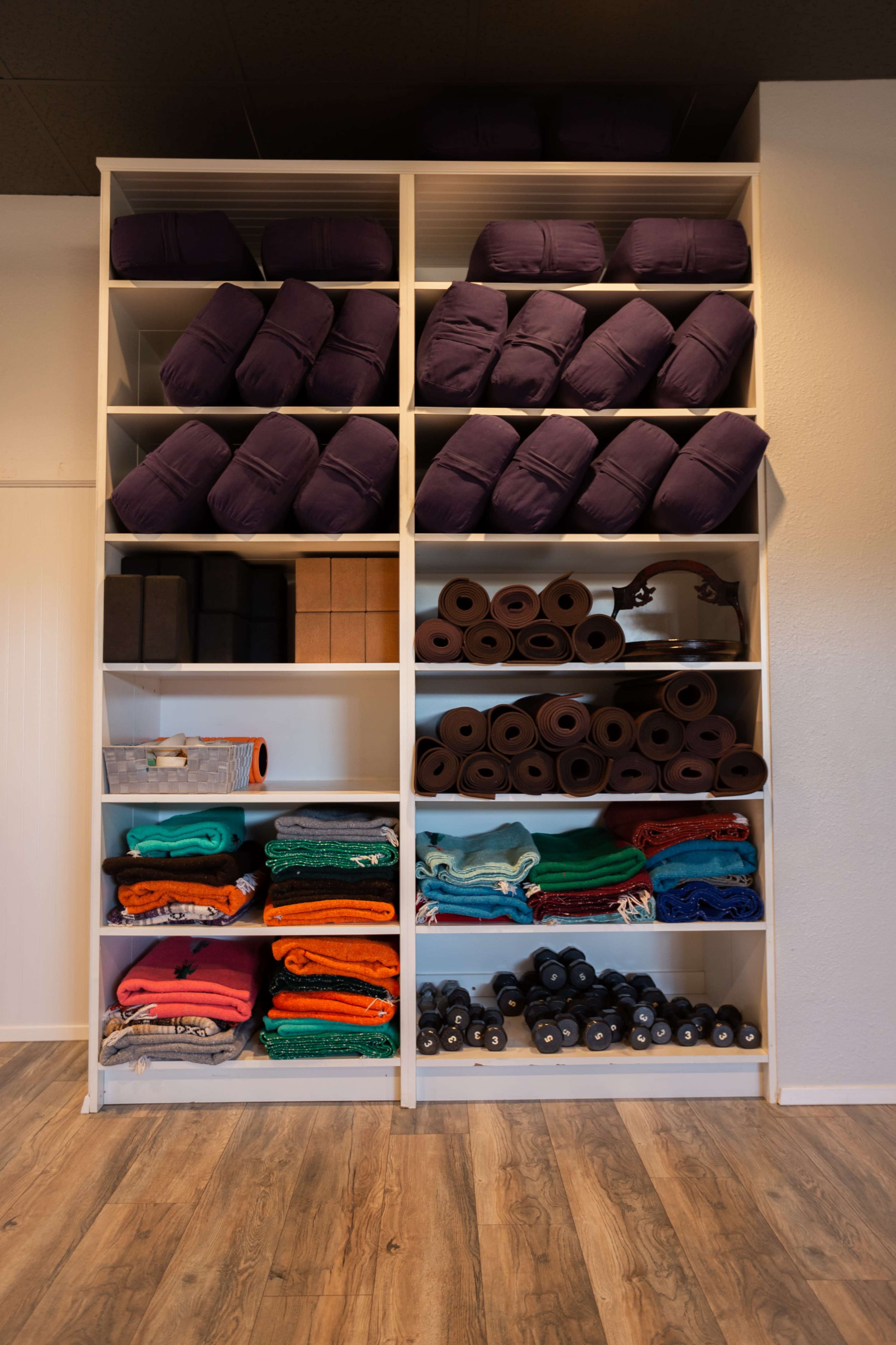 A neatly organized shelf displays stacks of purple yoga bolsters, blocks, mats, and colorful blankets, along with dumbbells in a fitness space.