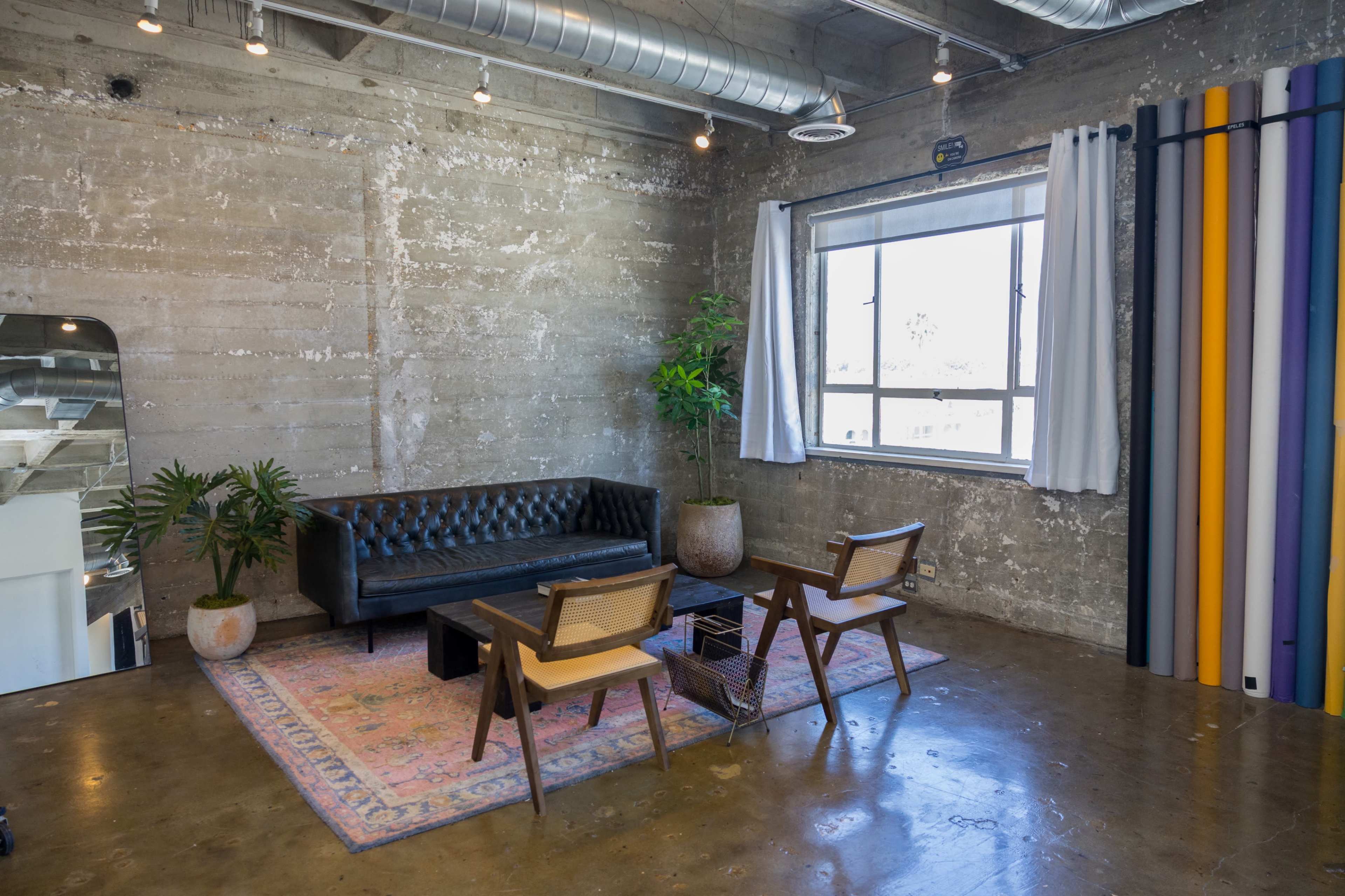 The image shows a modern, industrial-style interior featuring a black leather sofa, two wooden chairs, a decorative plant, and a colorful wall accent near a window.