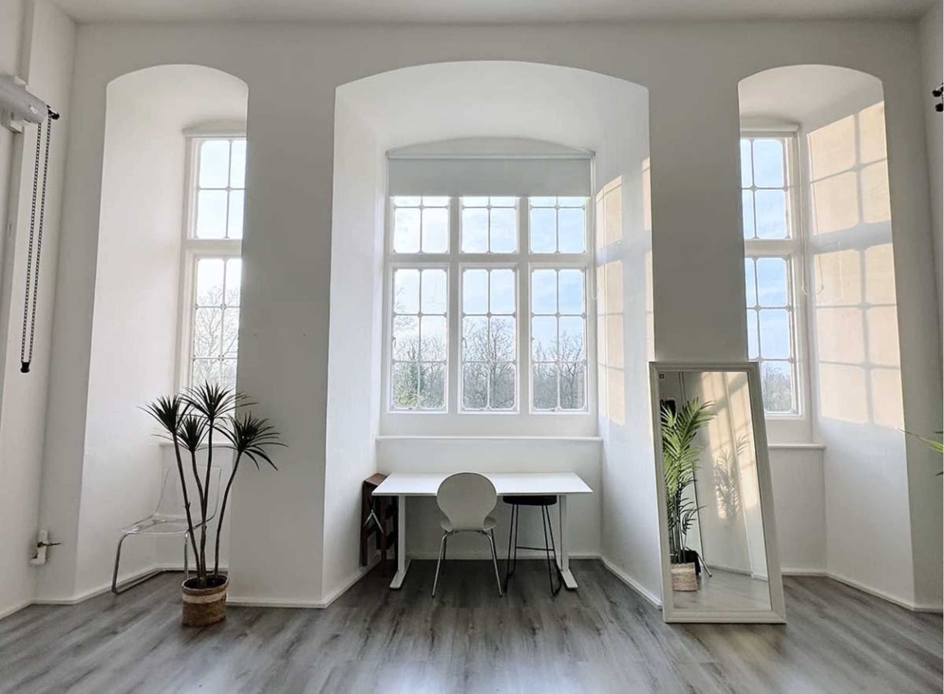 The image shows a bright, minimalist room with large windows, a small desk, a chair, and a tall mirror reflecting the light and plants.