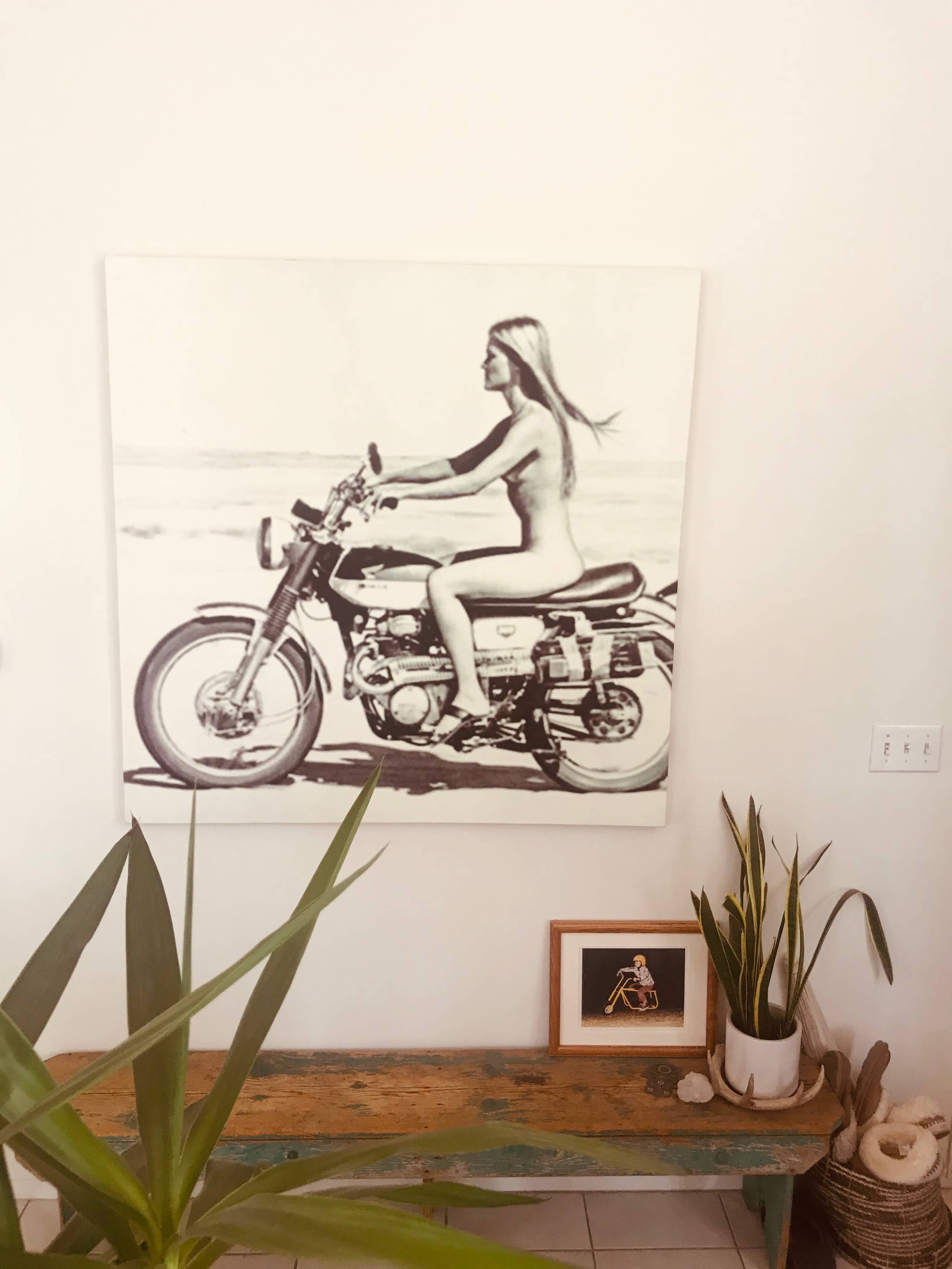 A black-and-white photograph of a woman riding a motorcycle is displayed on the wall above a wooden table with plants and a framed picture.