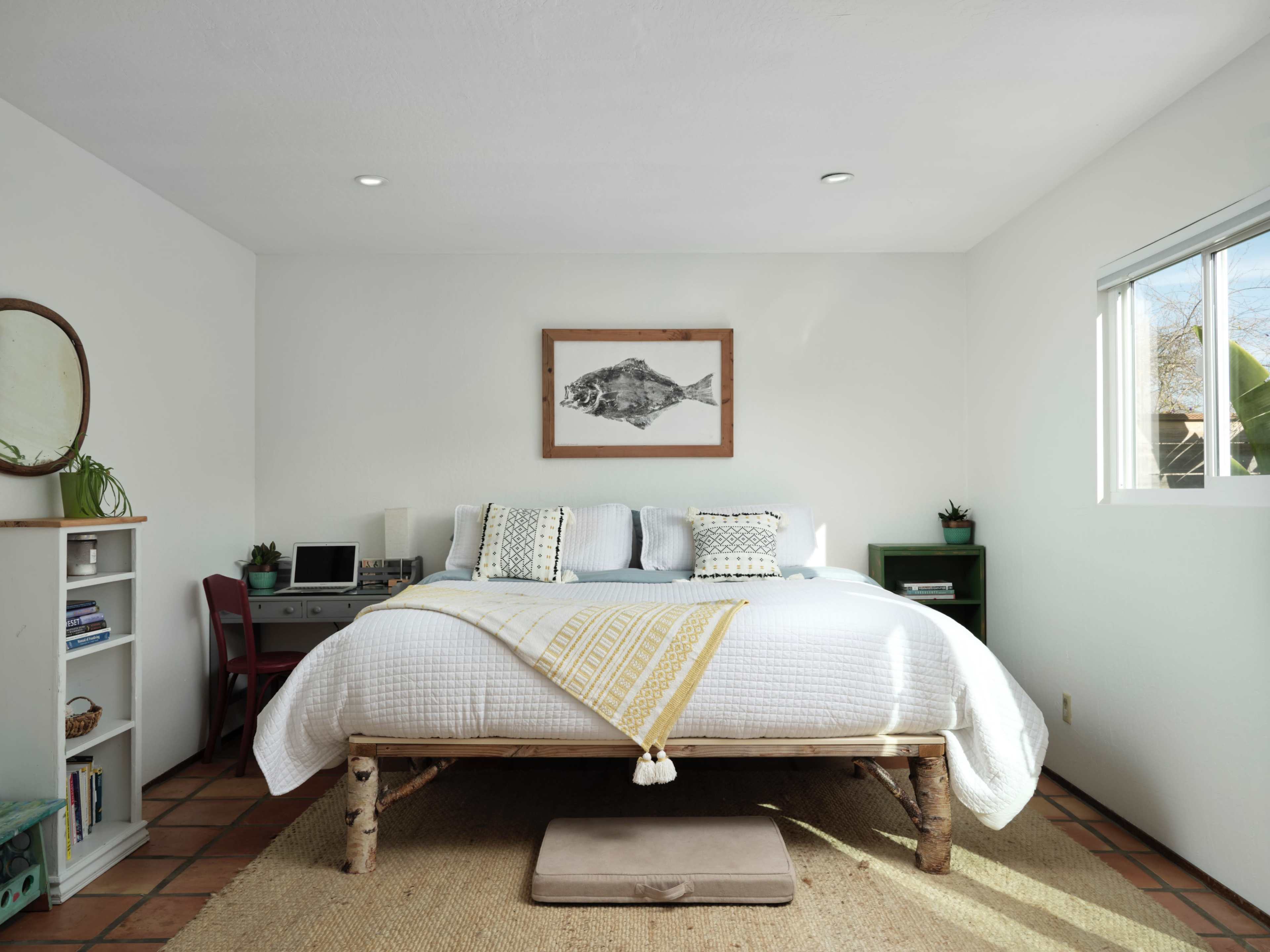 The bedroom features a bed with a white quilt and decorative pillows, a framed fish print on the wall, a small desk with a computer, and shelves holding books.
